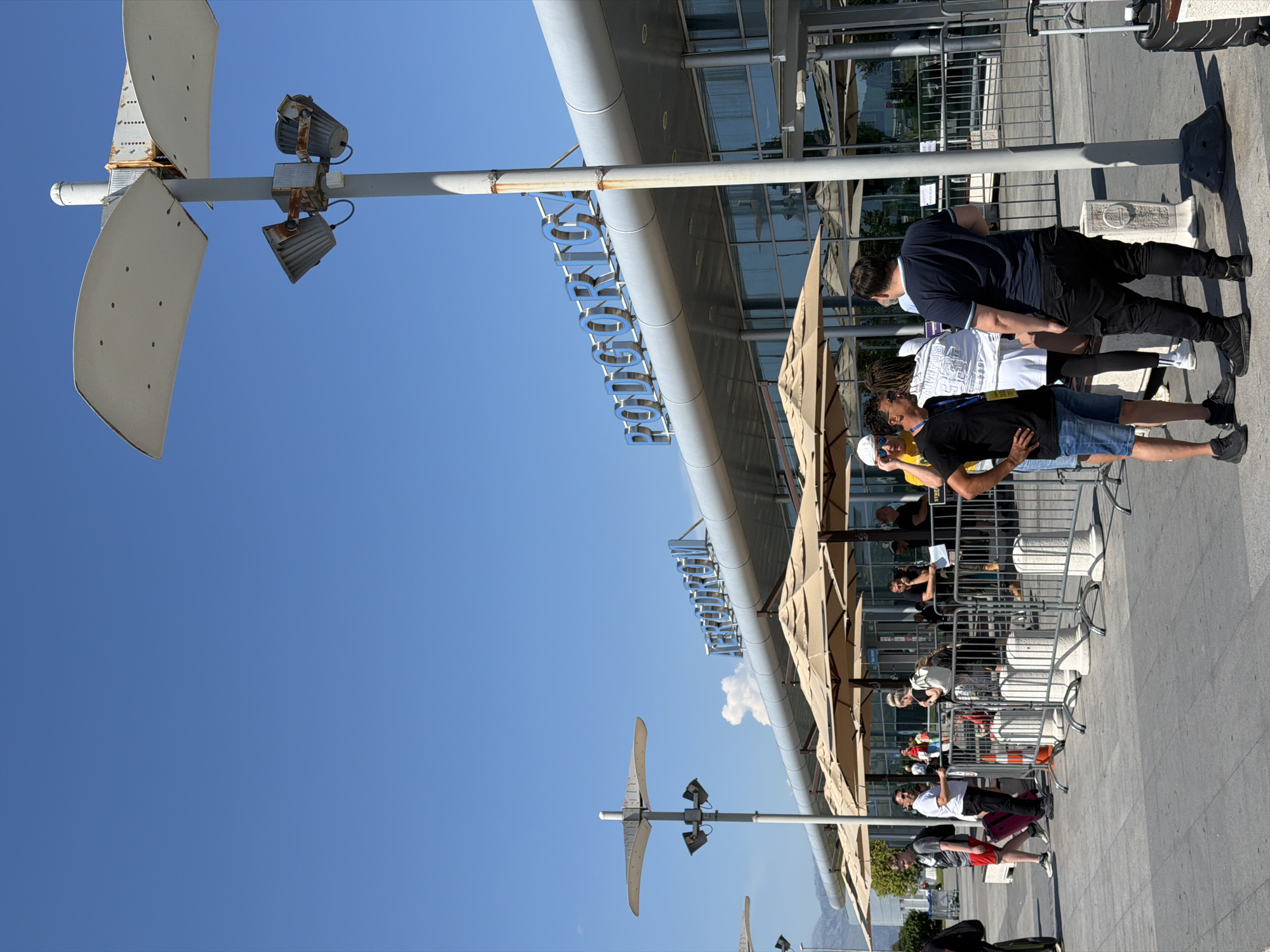 The image depicts the exterior of Podgorica Airport, located in Donja Cijevna, Montenegro. A bright, clear blue sky dominates the upper portion of the frame. The airport building, with its distinctive silver facade and large glass windows, is visible in the background. A large, illuminated sign spelling "PODGORICA" in blue capital letters is prominently displayed above the entrance.
In the foreground, a group of people are gathered outside the terminal, some standing in line behind temporary metal barriers. Several large, beige umbrellas provide shade over seating areas. Two prominent lampposts with modern, abstract designs and attached spotlights are present. One lamppost, closer to the viewer, has a white pole and appears to be made of metal.
The people in the scene are dressed in casual summer clothing. Some are seen with luggage, suggesting they are travelers. Interactions appear casual, with individuals talking amongst themselves or waiting in line. The ground is paved with gray concrete tiles. The overall atmosphere suggests a typical day at a regional airport.