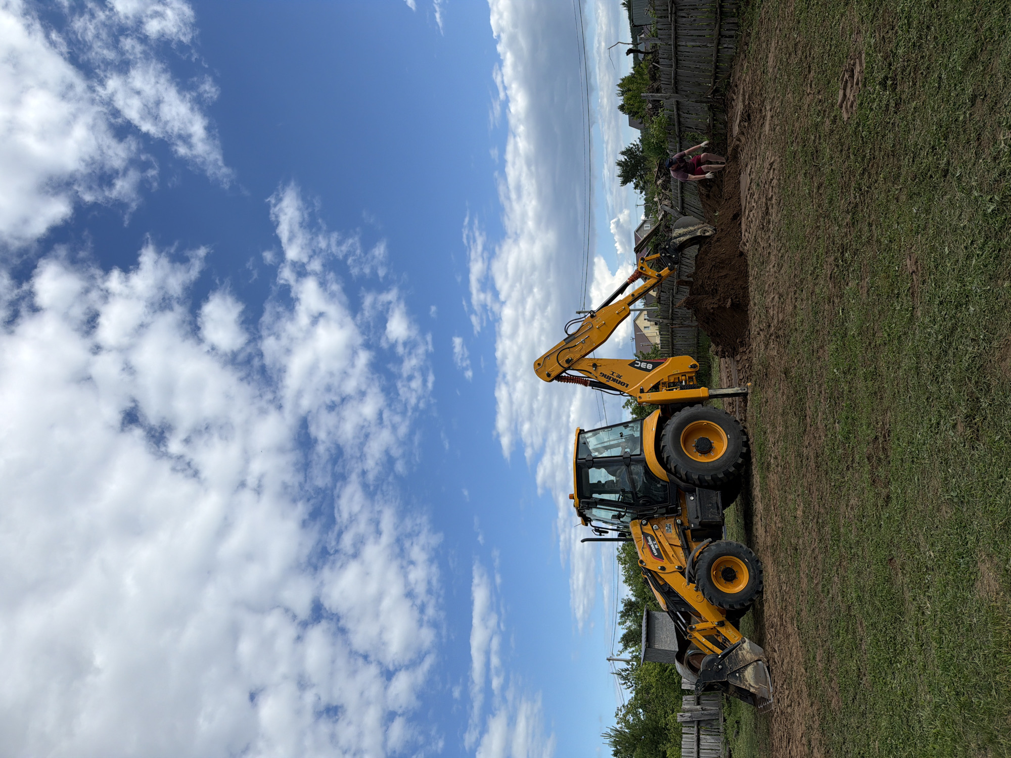 In Chaikovsky, Russia, on a sunny day with scattered clouds, a yellow backhoe loader is actively excavating dirt in a yard. The backhoe's arm is extended, and its bucket is in a pile of freshly dug earth, suggesting ongoing construction or landscaping work. A person, wearing a maroon t-shirt and red shorts with white gloves, is bending down near the pile of dirt, possibly assisting with the task or clearing debris. The yard is a mix of grass and disturbed soil, with a wooden fence running along the background. Several houses and trees are visible beyond the fence, indicating a residential area.