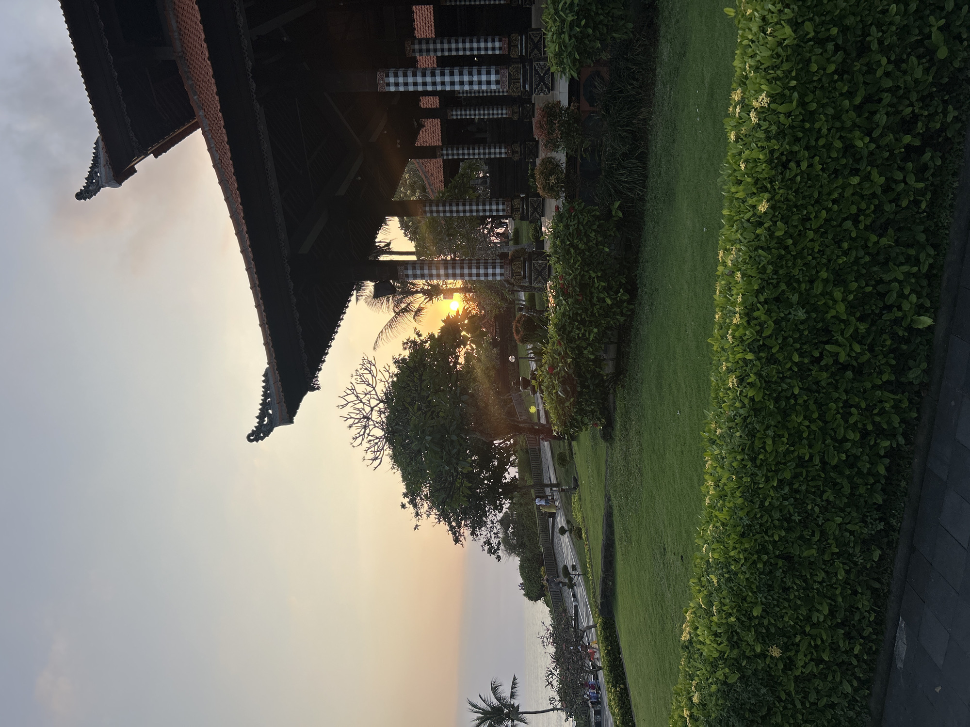 The image depicts a sunset scene at a location with traditional Balinese architecture, likely a temple or a resort, situated on a cliff overlooking the ocean. The sky is painted with warm hues of orange and yellow as the sun dips below the horizon, casting a soft glow on the landscape. A large, open-air pavilion with a tiled roof and intricately carved eaves dominates the right side of the frame. Its columns are adorned with black and white checkered fabric, a common motif in Balinese design. In the foreground, a meticulously manicured lawn slopes down towards the sea, bordered by vibrant green hedges. A pathway winds through the garden, with a few people strolling and enjoying the view. Palm trees and lush tropical vegetation add to the serene atmosphere. The ocean is calm, with gentle waves lapping at the shore below. While the image evokes a sense of peace and natural beauty, it is important to note that this scene is not located in the city of Kediri, Indonesia. The architectural style and coastal setting are characteristic of Bali, Indonesia, not Kediri, which is an inland city.