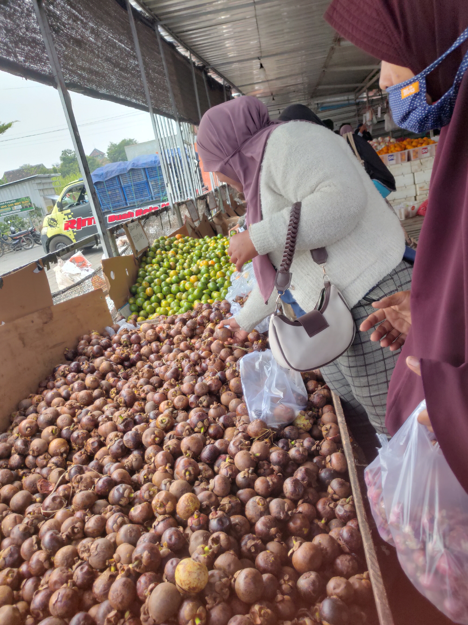The image captures a vibrant outdoor market scene in Ngasem, Indonesia, likely focusing on fresh produce. In the foreground, a large pile of dark-skinned mangosteens fills the frame, with a few yellow-tinged fruits interspersed. To the right, a person wearing a dark hijab and a blue polka-dot face mask, along with a burgundy garment, is holding a clear plastic bag filled with what appear to be red onions or shallots. Beside them, another person, wearing a light-colored sweater and a purple hijab, is reaching towards the mangosteens, holding a beige and brown handbag with a braided strap. In the mid-ground, a heap of green and yellow citrus fruits, possibly oranges or mandarins, is visible. Behind the produce stalls, a yellow truck with red and black lettering is parked, and a blue tarp-covered truck can be seen in the distance. The market is covered by a corrugated metal roof supported by metal poles, with a woven matting overhead creating shade. The overall impression is one of a bustling local market, showcasing the abundance of fresh fruits and vegetables.