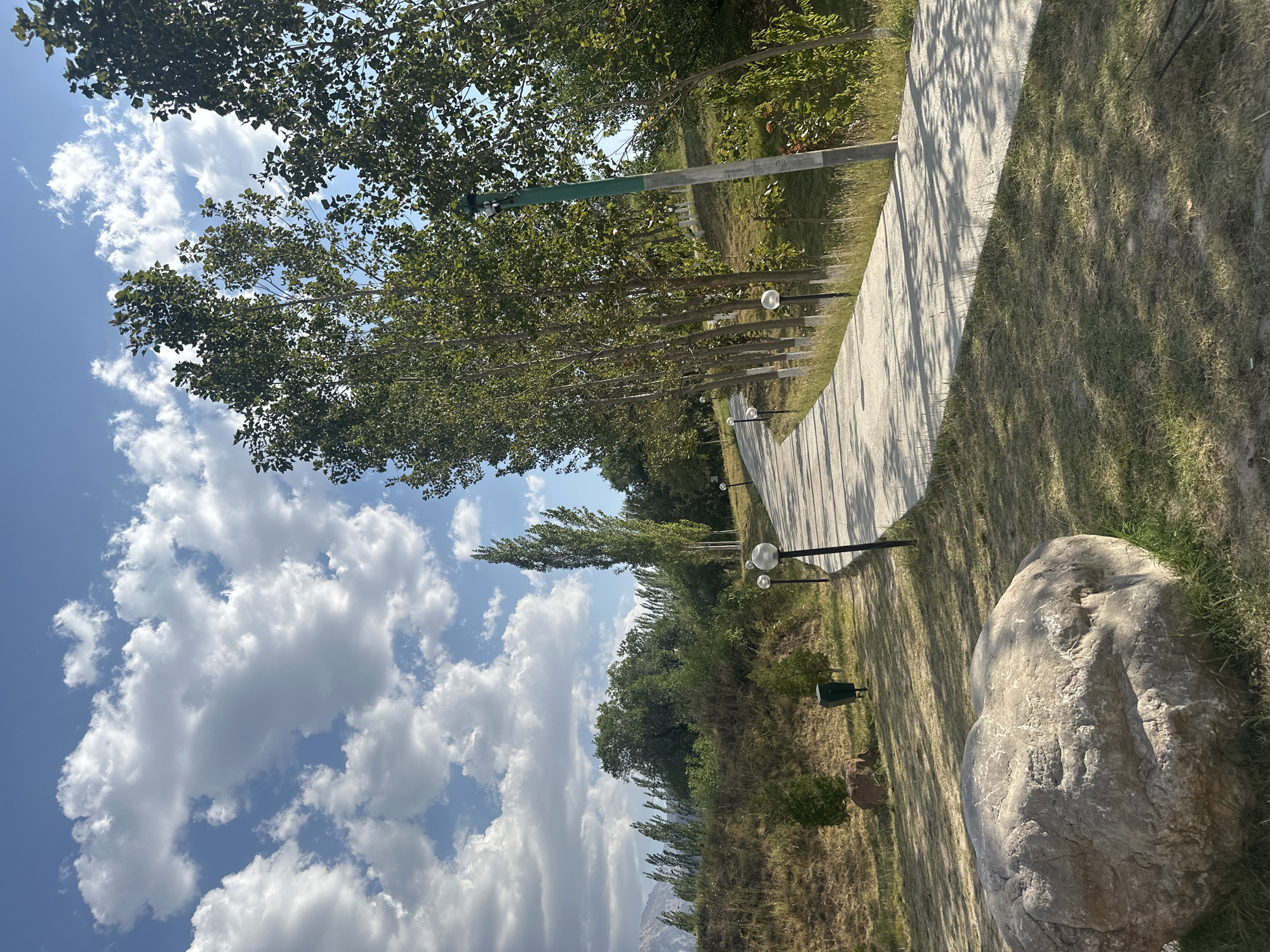 The image depicts a scenic view of a paved walking path winding through a park-like setting, likely in the city of Yusuphana, Uzbekistan. The path is lined with evenly spaced trees, casting dappled shadows on the ground, and several modern-style lampposts. To the left of the path, a grassy slope descends, sparsely dotted with small trees and a large boulder in the foreground. Further up the slope, a trash receptacle and a small animal, possibly a deer, are visible. The background features a dense line of trees, with a hazy mountainous landscape in the distance. The sky is a vibrant blue, adorned with fluffy white clouds, suggesting fair weather. The overall impression is one of a well-maintained outdoor recreational area on a clear day.