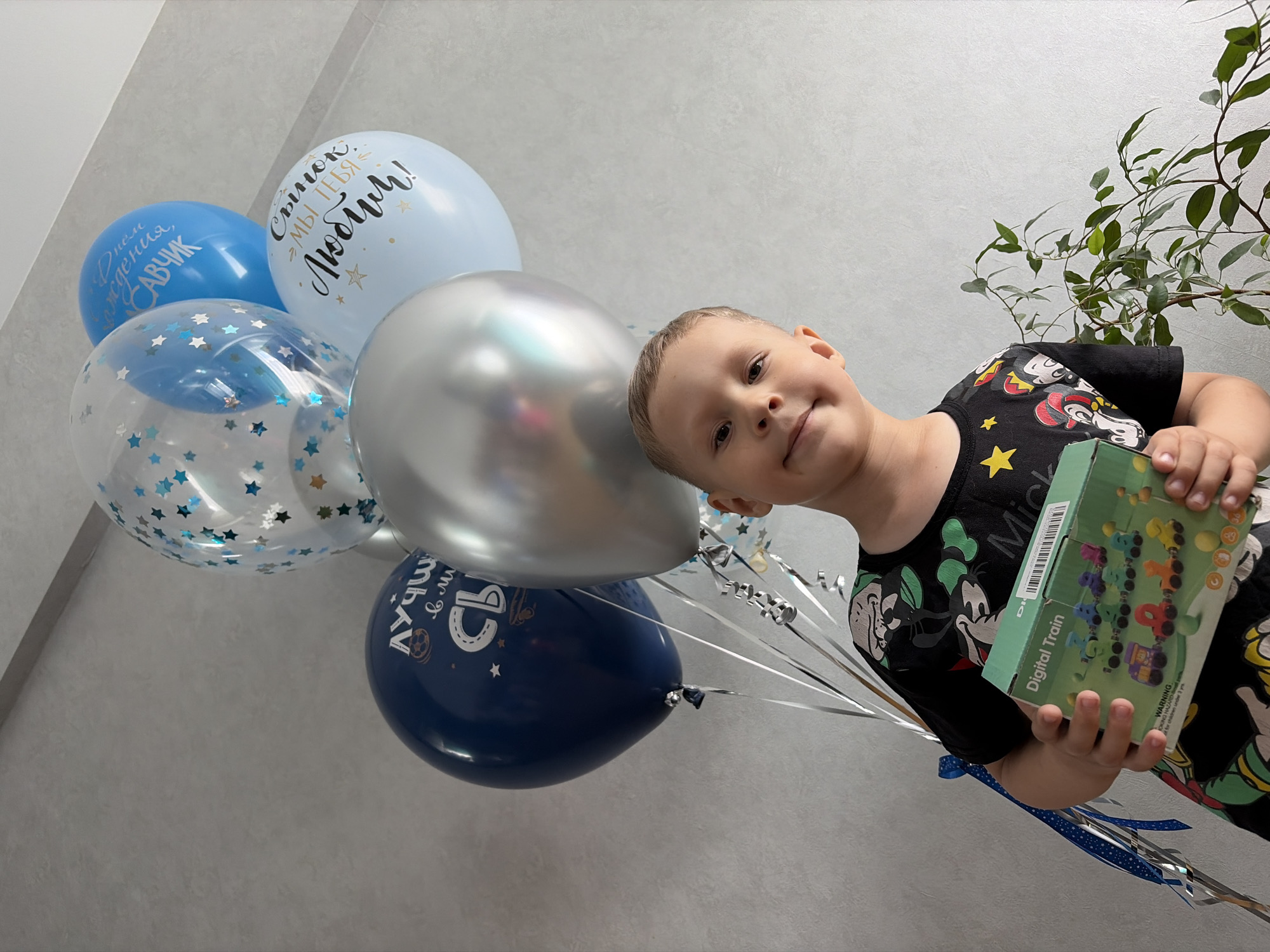 A young Caucasian boy, approximately 5-7 years old, stands in front of a grey textured wall, holding a gift box. He is wearing a black t-shirt with cartoon characters. The boy is smiling. In the background, there are several balloons: one clear balloon filled with star confetti, a light blue balloon with text, a navy blue balloon with text and a soccer ball graphic, a silver balloon, and another light blue balloon with text. There is also a green potted plant to the right of the boy. The scene appears to be indoors, likely a residential setting. The image does not contain enough information to determine its specific location within Chaikovsky, Russia.