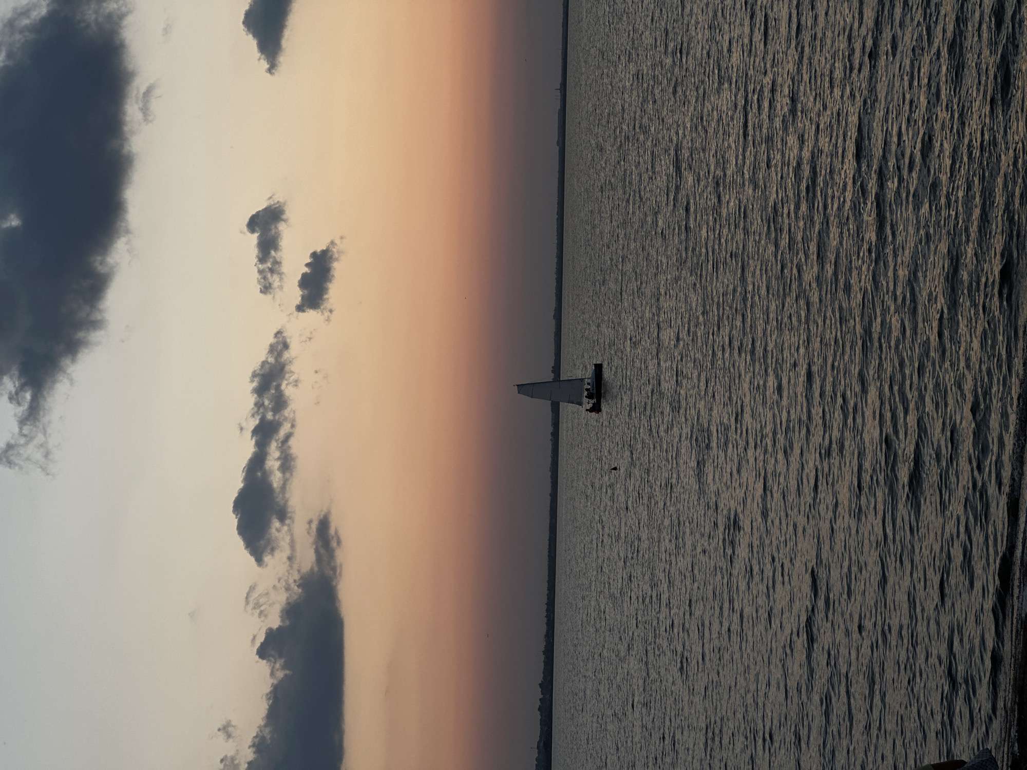 A sailboat with a white sail navigates calm water under a twilight sky. Dark, puffy clouds are scattered across the upper portion of the sky, which transitions from a light grey at the top to a soft orange and then a deep purple towards the horizon. The horizon line is marked by a dark, silhouetted landmass. The water is a deep, dark blue with subtle ripples reflecting the muted light. A few figures can be seen on the deck of the sailboat. The scene appears to be a tranquil evening on a large body of water, possibly a lake or sea, with the distinctive silhouette of the Kokshetau city skyline faintly visible in the distance across the water.