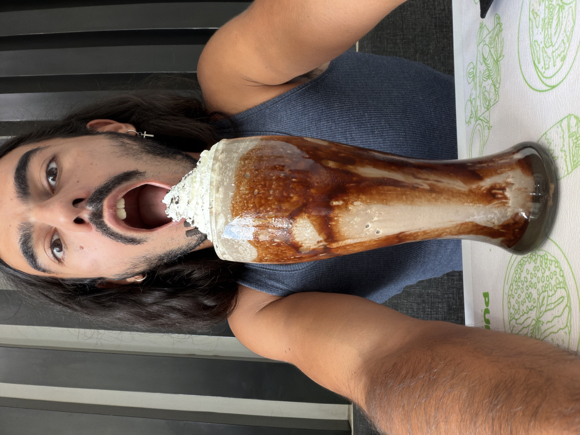 A young adult with long dark hair and a mustache is leaning towards the camera with their mouth open as if to take a bite of a large chocolate milkshake. The milkshake is in a tall, clear glass with whipped cream and chocolate syrup drizzled down the sides. The person is wearing a dark blue tank top. The background appears to be an indoor setting with vertical dark blinds. The scene is set on a table with a white placemat featuring a green line drawing of food items. The image is a close-up, capturing the person's face and the milkshake from the chest up.