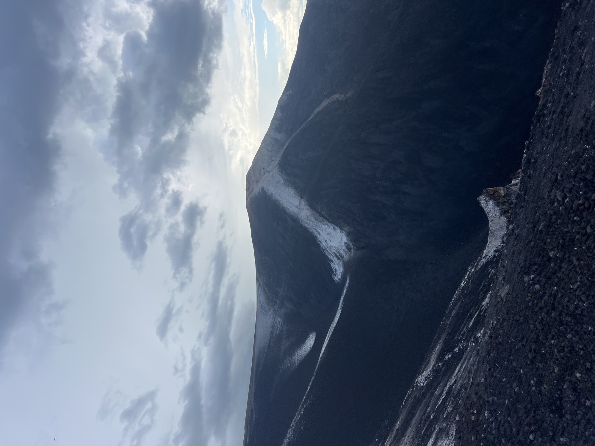 The image depicts a steep, dark, volcanic landscape under a cloudy sky. The primary subject is a mountain, likely a volcano, with a dark, rocky surface. A prominent feature is a lighter-colored streak running down the side of the mountain, possibly indicating a path or a geological formation. The foreground is also composed of dark, loose volcanic rock. In the distance, the sky is filled with dramatic, heavy clouds, with patches of brighter sky and sunlight breaking through. The overall impression is one of a rugged and imposing natural environment. The scene appears to be set during the daytime, but the cloudy conditions suggest it could be overcast or approaching stormy weather. The specific location, Rota, Nicaragua, is known for its volcanic activity and dramatic landscapes, which aligns with the visual cues in the image. There is no visible text in the image.