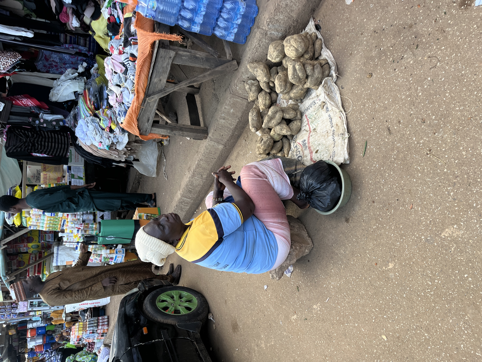 This image depicts an outdoor market scene in Bukuru, Nigeria. A woman, dressed in a white beanie, a blue and yellow shirt, and pink and white patterned pants, is seated on a rock. She is positioned in the foreground, looking to the right with her hands clasped. In front of her is a black trash bag in a green bowl, and to her right, a pile of yams is spread on a white sack.
Behind the woman, a man in a brown traditional outfit and a patterned cap stands near a green receptacle and a table laden with various goods. Further back, other individuals can be seen amidst stalls displaying a wide array of items, including food products, clothing, and bottled water. A black car with a distinctive green wheel is parked on the left side of the frame. The ground is paved with rough asphalt, and the overall environment suggests a busy commercial area.