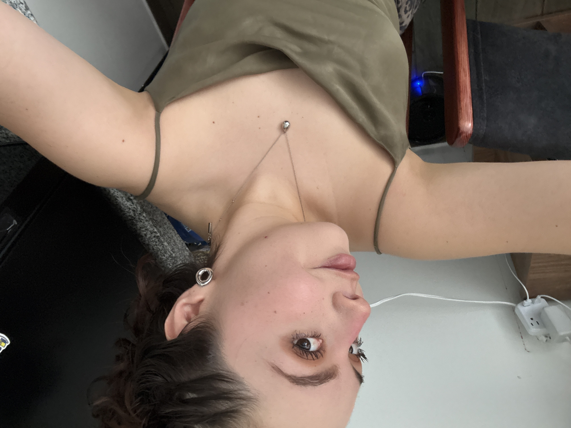 A woman with brown curly hair and gauged ears is seen wearing a pale green tank top with thin straps and a silver necklace. The image is oriented upside down. She is looking towards the camera with brown eyes and fair skin. A white power strip with cords is visible on a white wall in the background. Part of a black surface, possibly a table or countertop, is visible on the left. A chair with dark upholstery and a wooden frame is partially visible on the right. The location of the image is unknown and no details suggest the city of Mexico City.