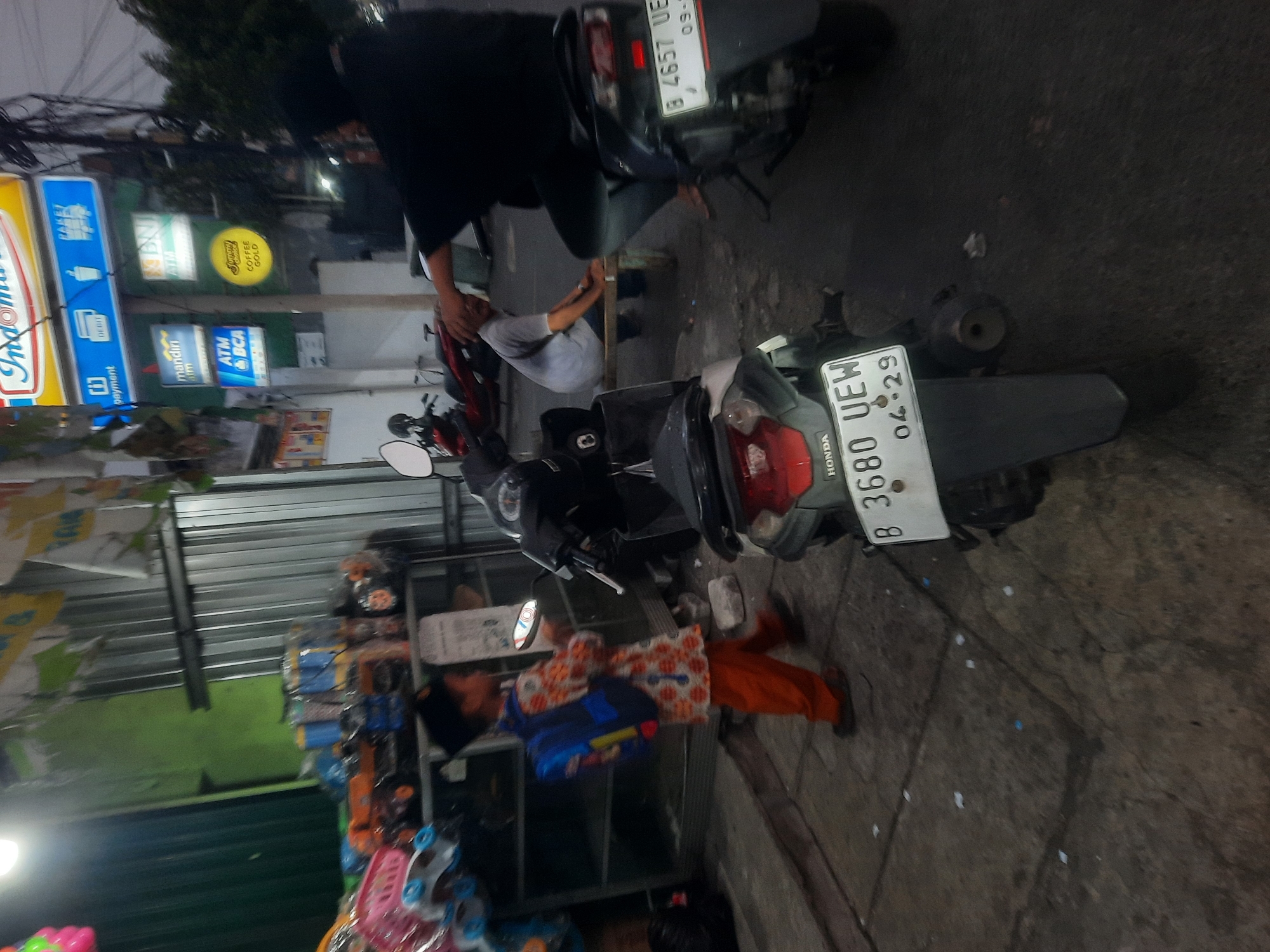 The image depicts a street scene in North Jakarta, Indonesia, likely in a commercial area with multiple small businesses. In the foreground, a young boy wearing an orange and white patterned shirt and orange pants stands with a blue backpack. He is holding a small, circular object, possibly a toy or a device. To his right, a black motorcycle is parked, its license plate clearly visible: B 4657 UEW 04-29. Further back, another motorcycle, red and black, is visible.
In the middle ground, a person wearing a dark shirt is seated on a bench, facing away from the viewer. Behind them, several signs are visible, including an "Indomaret" logo, indicating a convenience store chain. Other signs suggest banking services, with logos for "Mandiri ATM" and "ATM BCA". The environment appears to be an urban street with corrugated metal structures, possibly part of storefronts or stalls. The lighting suggests it is either early morning or late evening. Scattered on the ground are small pieces of white debris.