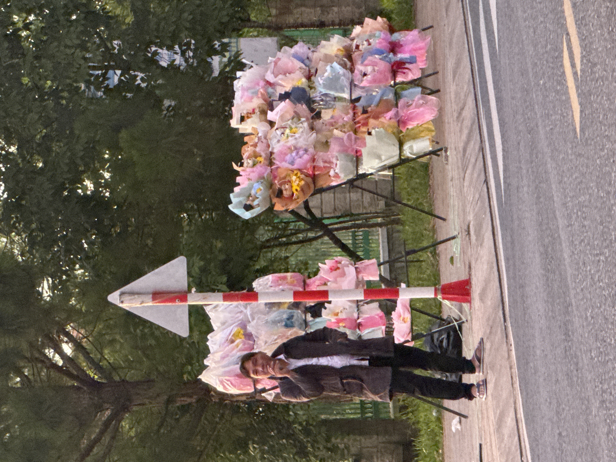 A man is standing next to a flower stand in Da Lat, Vietnam. He is wearing a dark coat over a white shirt and dark pants, with sandals on his feet. He is holding a cigarette to his mouth. The flower stand is filled with bouquets of artificial flowers wrapped in plastic, in various pastel colors like pink, purple, and blue. A red and white striped pole with a triangular sign is positioned in front of the stand. Behind the stand, there are trees and a low fence. The scene appears to be on a sidewalk next to a road.