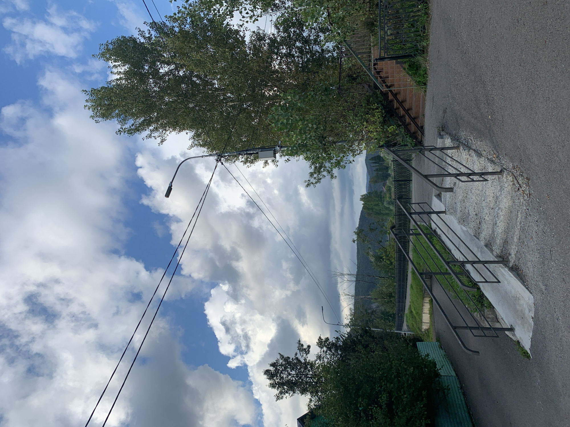 The image depicts an outdoor scene with a cloudy sky overhead. In the foreground, a paved pathway leads to a concrete stairway with metal railings. To the left of the pathway, a green fence borders an area with dense foliage. A street light is visible on a pole to the right of the stairway. In the background, more trees and a distant hillside are visible under the expansive sky. The scene appears to be in a town or city, possibly with some residential structures partially visible on the left.