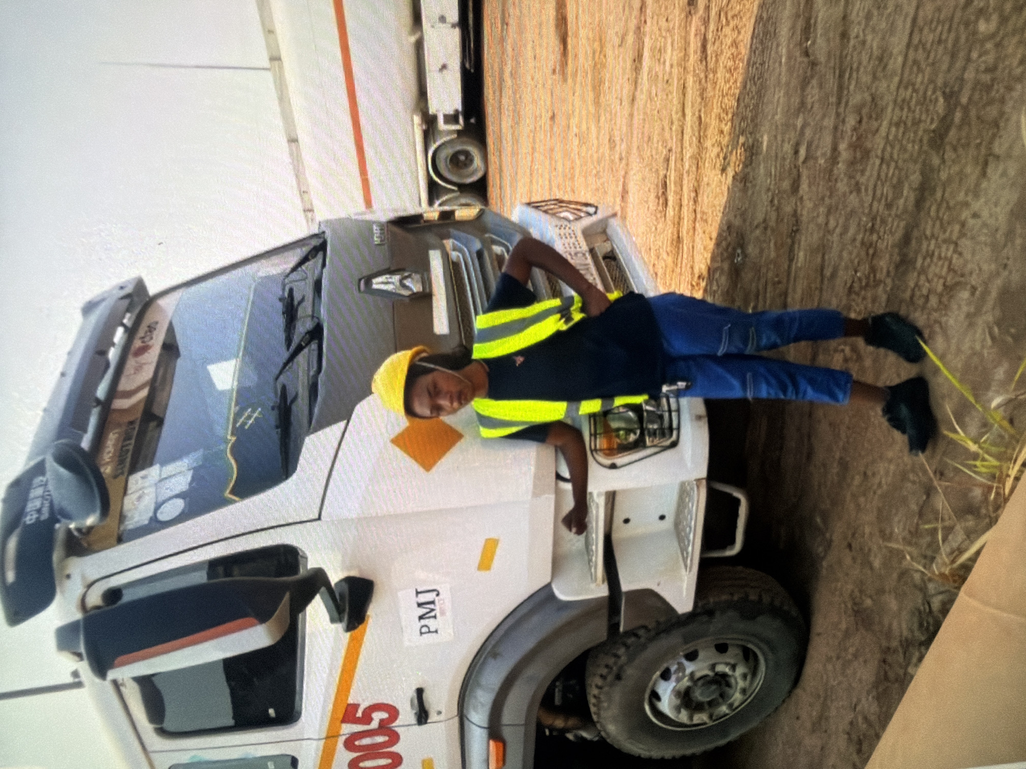 A woman wearing a yellow hard hat and a neon yellow safety vest stands in front of a white semi-truck with the number "005" visible in red lettering on the side. She has her hands on her hips and is looking towards the camera. The truck appears to be a tanker truck. The ground is dirt and there are some sparse patches of grass. In the background, another tanker truck is visible. The overall environment suggests an industrial or construction setting.