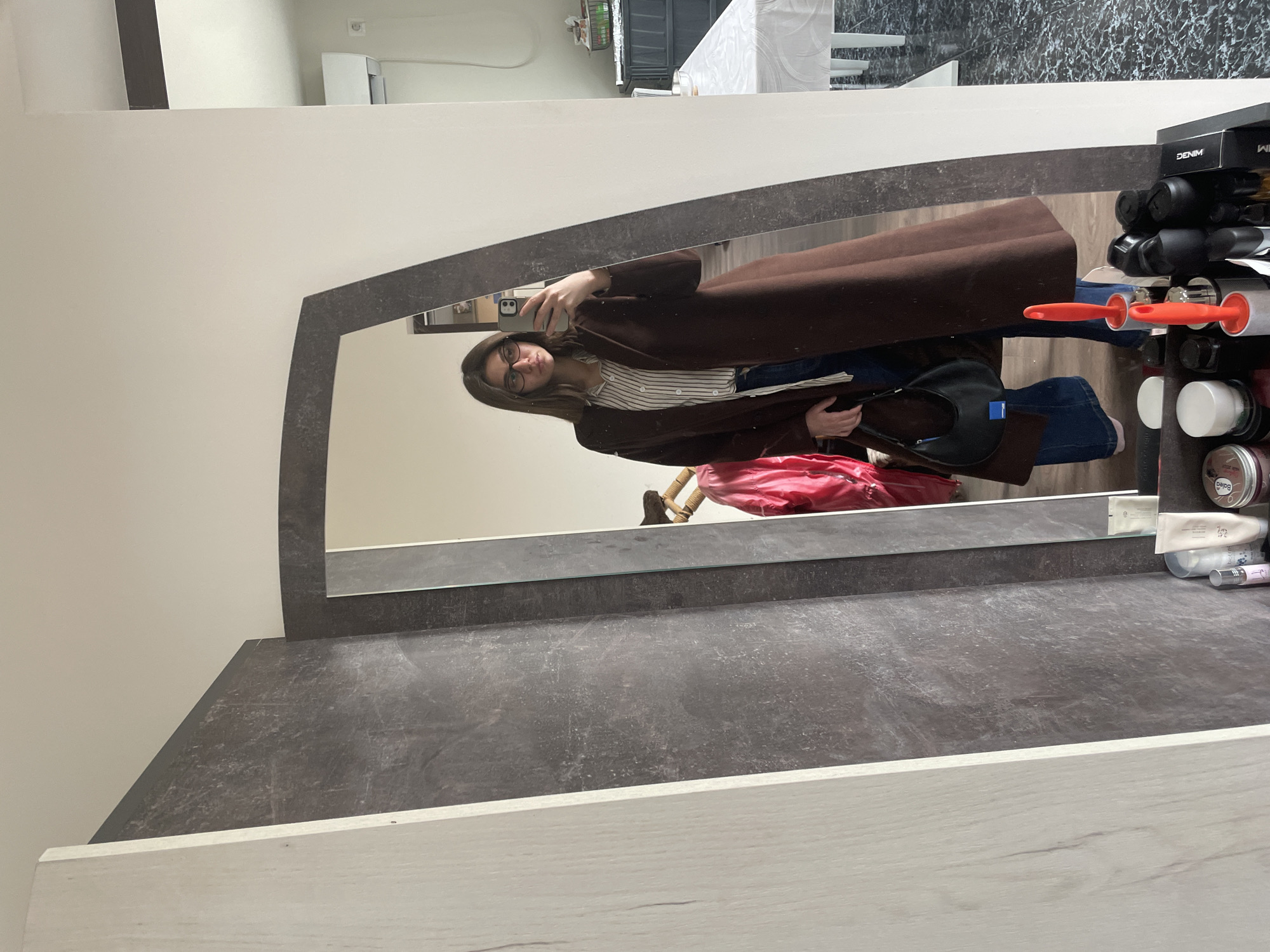 The image shows a person in a mirror, likely taking a selfie. The person is wearing glasses, a striped shirt, jeans, and a brown coat. A black purse is visible. The mirror is mounted on a gray marble-look surface, possibly part of a vanity or shelf. Household items, including cleaning supplies, are arranged near the edge of the surface. The room appears to be a residential interior. The presence of a reflection of a table with a white tablecloth, and a modern staircase visible in the background, suggests the room is a hallway or entryway, rather than a bathroom. The location in the city of Sofia, Bulgaria, cannot be forensically confirmed from the image alone.