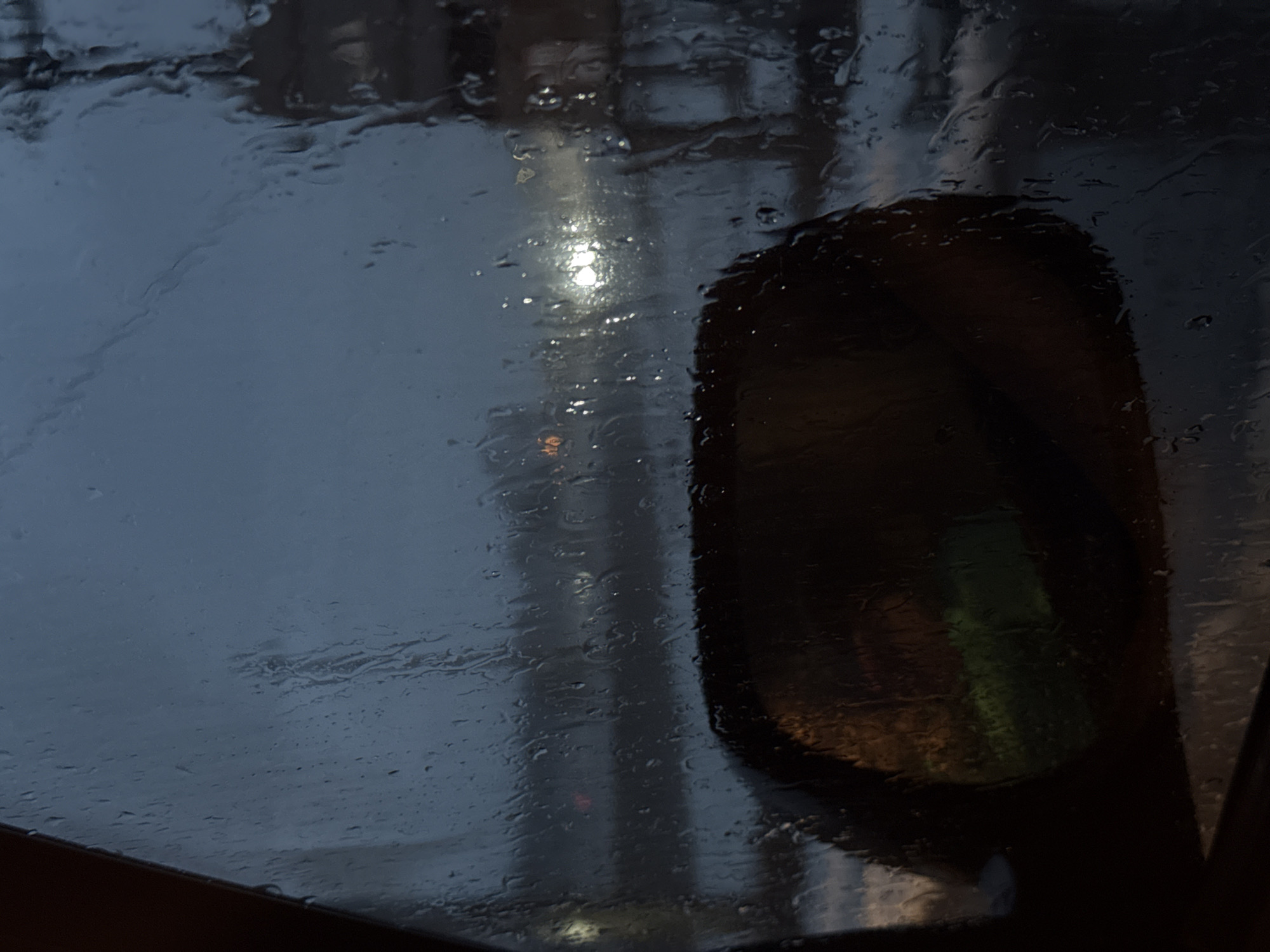 The image depicts a rain-streaked window, likely of a vehicle, with a side mirror visible on the right. The view through the window is obscured by the rain, but a blurred light source can be seen in the distance. A faint green and brown image is reflected in the side mirror. There is no information in the image indicating that the location is Yazlova, Russia.