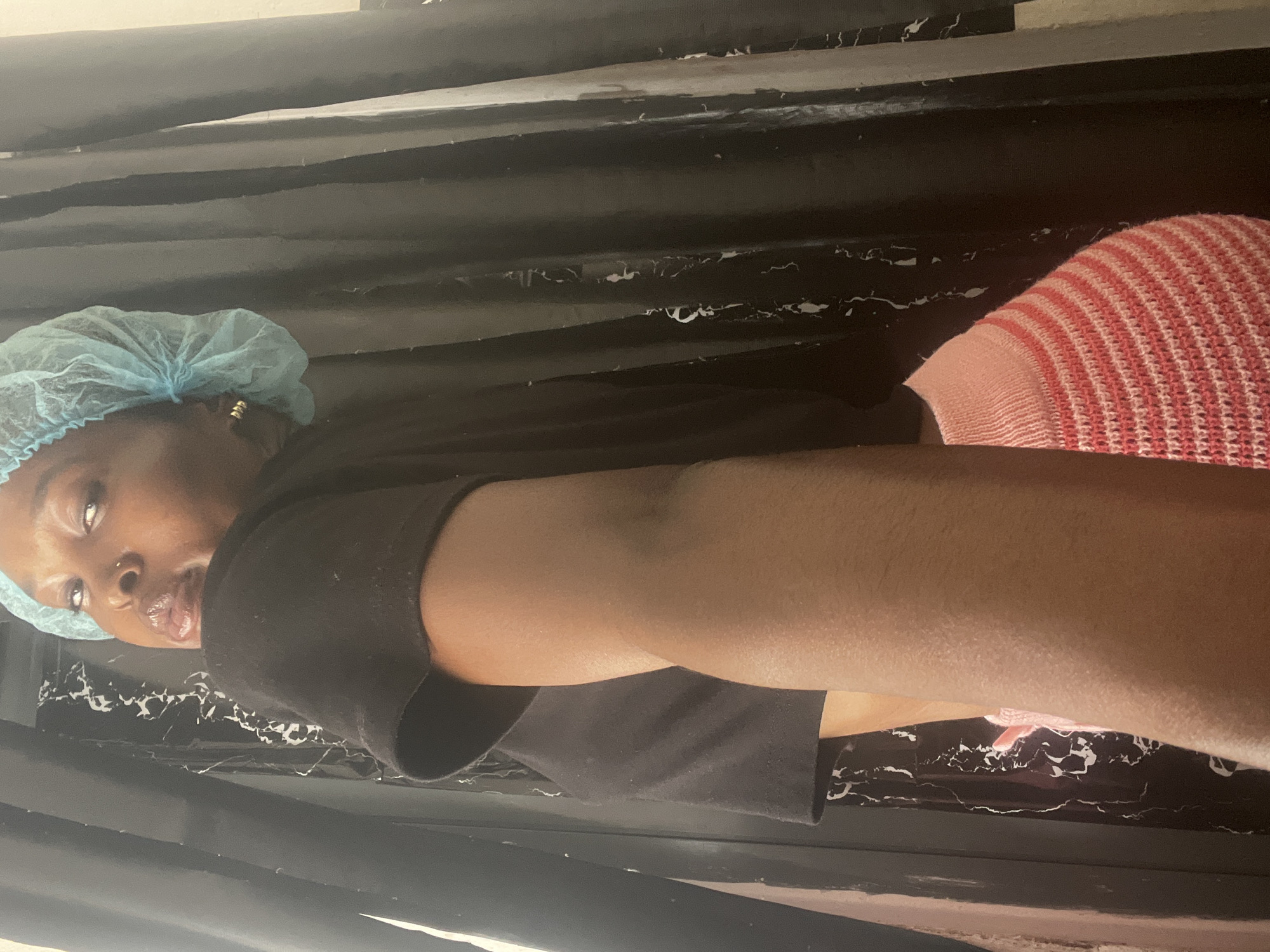 A woman with dark skin wearing a light blue surgical cap, a black t-shirt, and a pink and white striped sweater is lying on her side on a black surface that appears to be marble. The surface could be a counter or a table. The photo's location is specified as Lekki, Nigeria.
