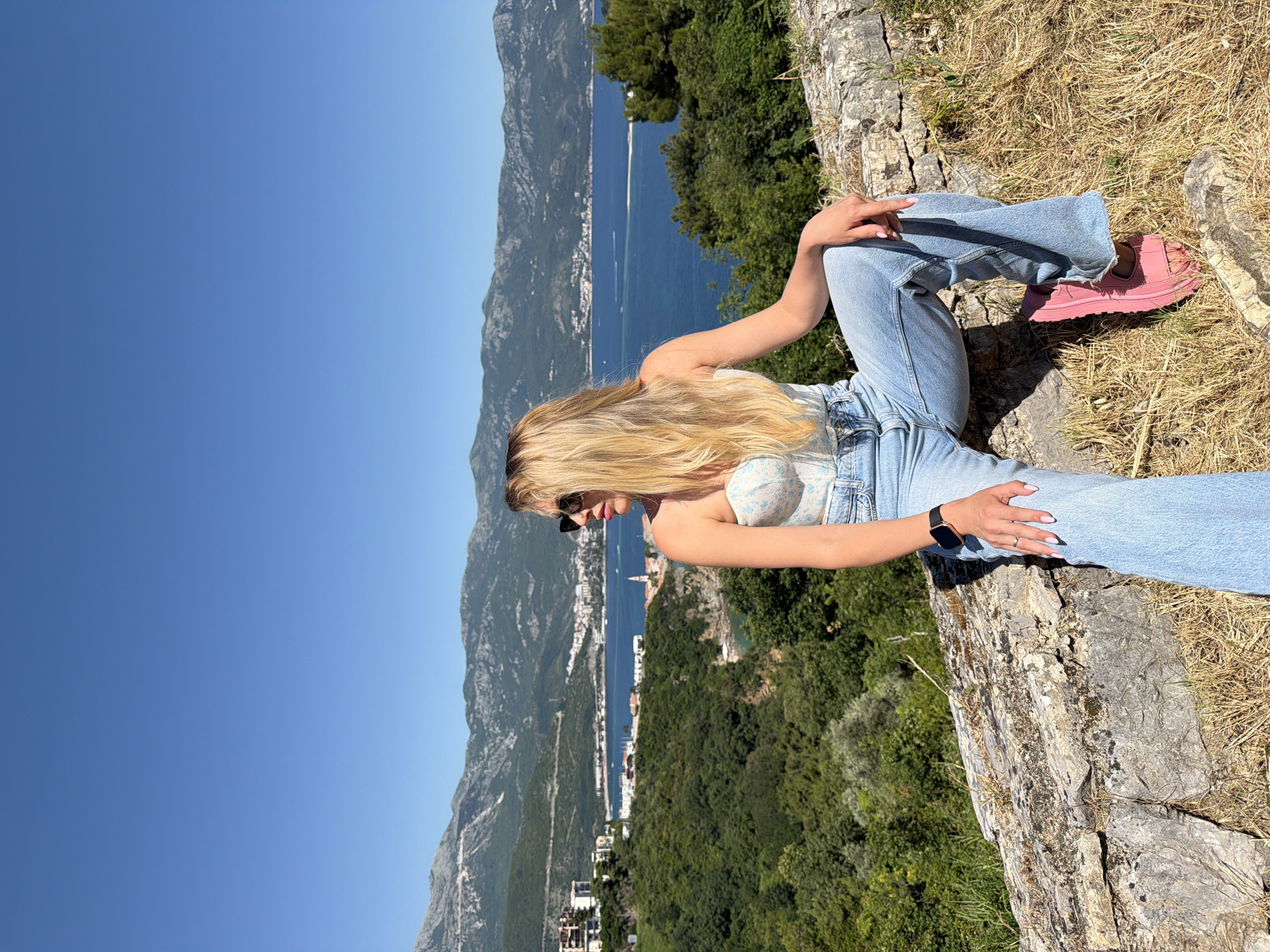 A young woman with long blonde hair and sunglasses sits sideways on a weathered stone wall, facing right. She is wearing a white, patterned strapless top, light blue wide-leg jeans, and bright pink slides. A smartwatch is visible on her left wrist, and a ring on her left index finger. In the immediate foreground are dry grass and scattered rocks. Beyond the wall, a verdant hillside slopes down towards the Adriatic Sea. Distant buildings and a church spire of Budva are visible along the coastline. The wide blue expanse of the sea features a visible sandbar or landmass and boats. Majestic mountains with varying green and barren slopes form the backdrop under a clear, cloudless blue sky, characteristic of the Montenegro landscape surrounding Budva. The scene is brightly illuminated by direct sunlight, casting distinct shadows.