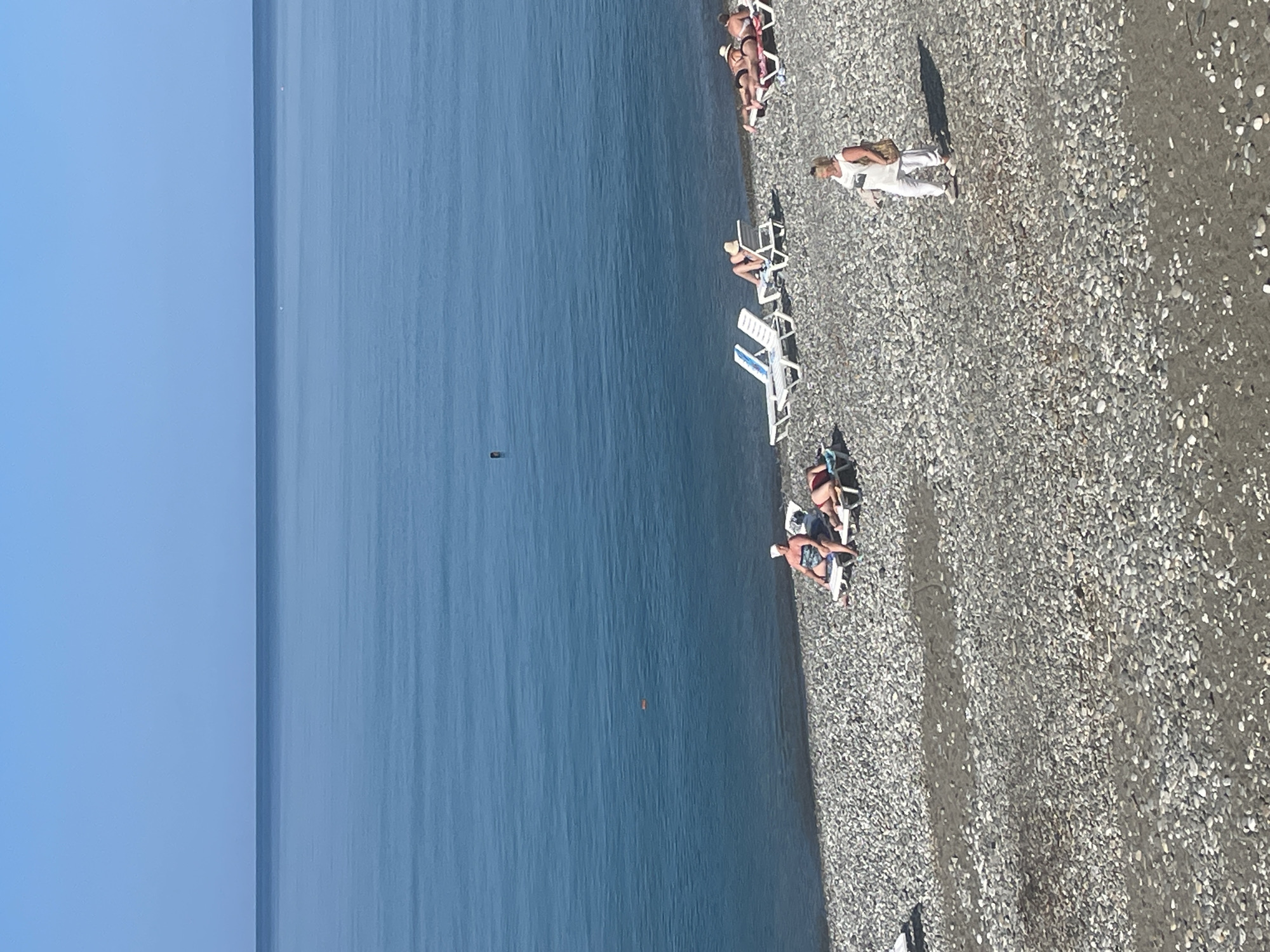 The image captures a sunny day at a pebble beach in Giachrypsh, Abkhazia, with the vast, calm blue sea stretching to the horizon under a clear sky. The foreground is dominated by a dense covering of smooth, gray pebbles. Several lounge chairs are placed on the beach, some occupied by sunbathers. In the mid-ground, a woman in a white top and pants walks across the pebbles, carrying a bag over her shoulder. Further out in the water, a dark rectangular object, possibly a buoy or marker, is visible. Another small, orange object floats in the water closer to the shore. The overall scene depicts a peaceful day at the seaside.