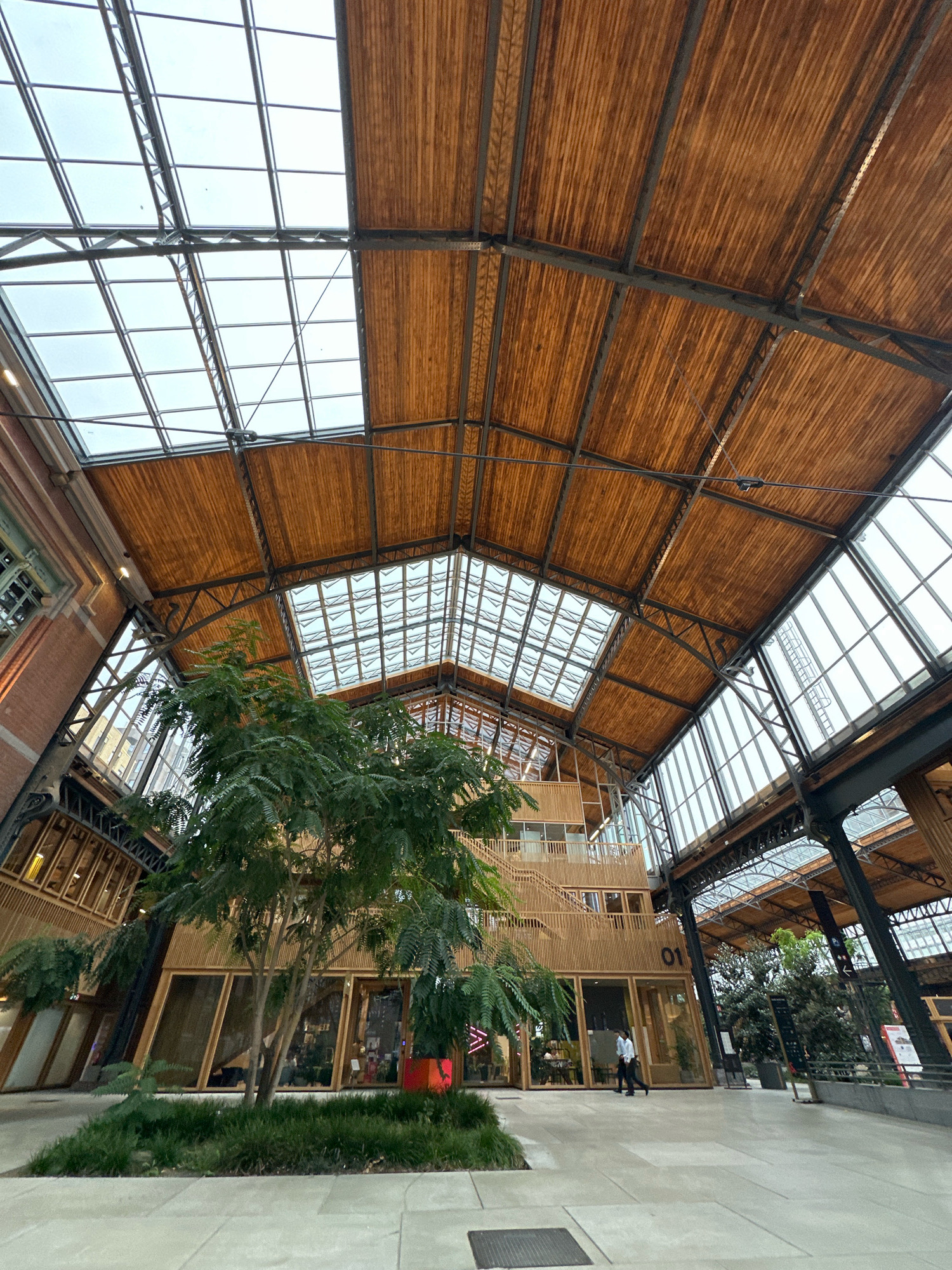 The image depicts the interior of a spacious, modern building with a high, arched ceiling constructed from exposed wooden beams and glass panels. The architecture features a prominent central atrium with a large tree and green foliage. A multi-story wooden structure with balconies and large glass windows is visible within the atrium, appearing to house commercial or office spaces. Several individuals are present, including one person walking in the foreground and others visible through the windows of the wooden structure. The overall ambiance suggests a contemporary, well-lit indoor public space, possibly a shopping center, office complex, or cultural venue. The location is identified as Molenbeek-Saint-Jean - Sint-Jans-Molenbeek, Belgium.
