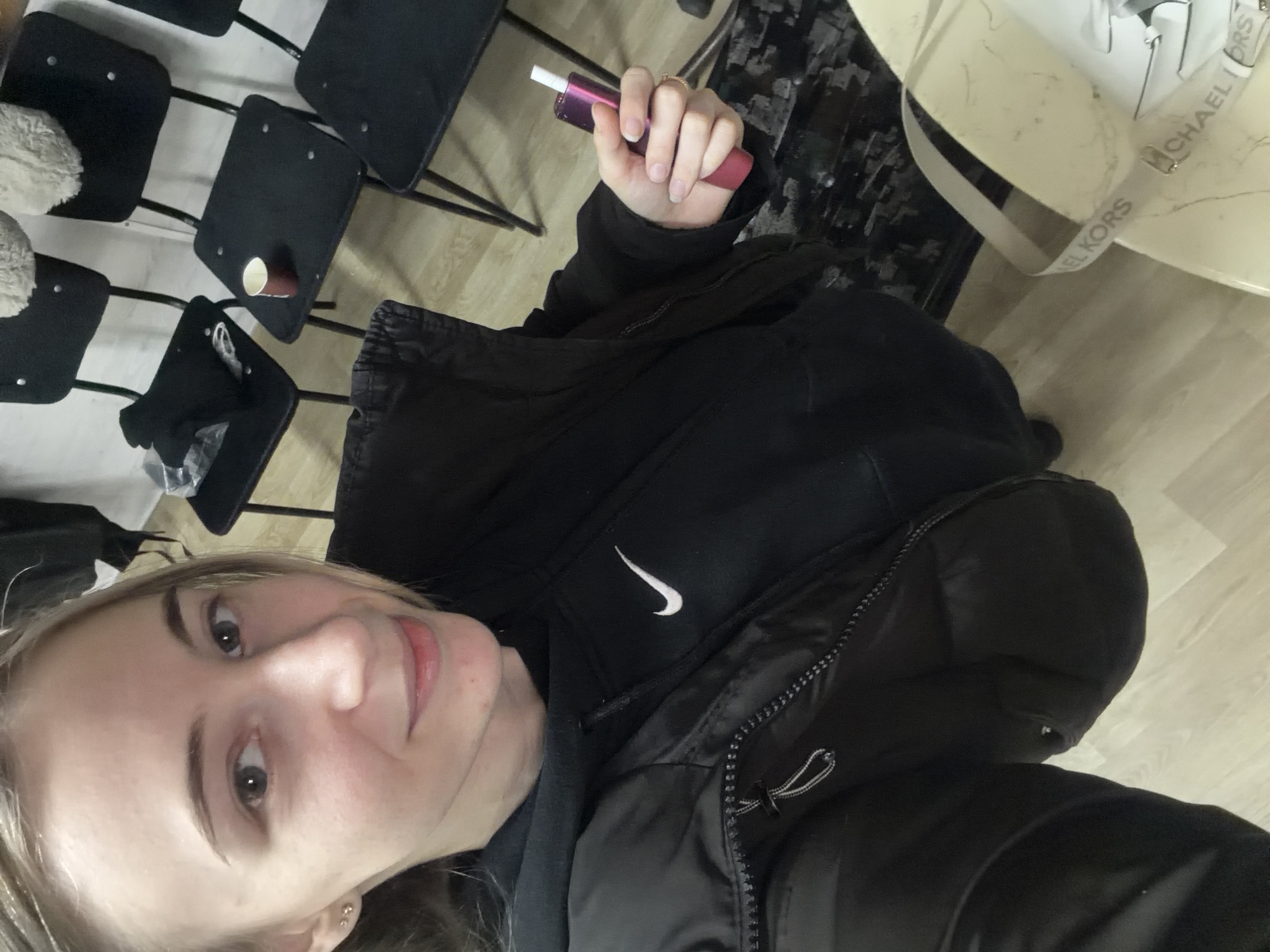 A woman is taking a selfie. She is wearing a black jacket with a white Nike logo. She is holding a purple and pink cylindrical object with a white tip. There are black chairs visible in the background. A Michael Kors handbag is visible on a table in the background. The environment suggests an indoor setting. No specific location in Plovdiv, Bulgaria is evident.