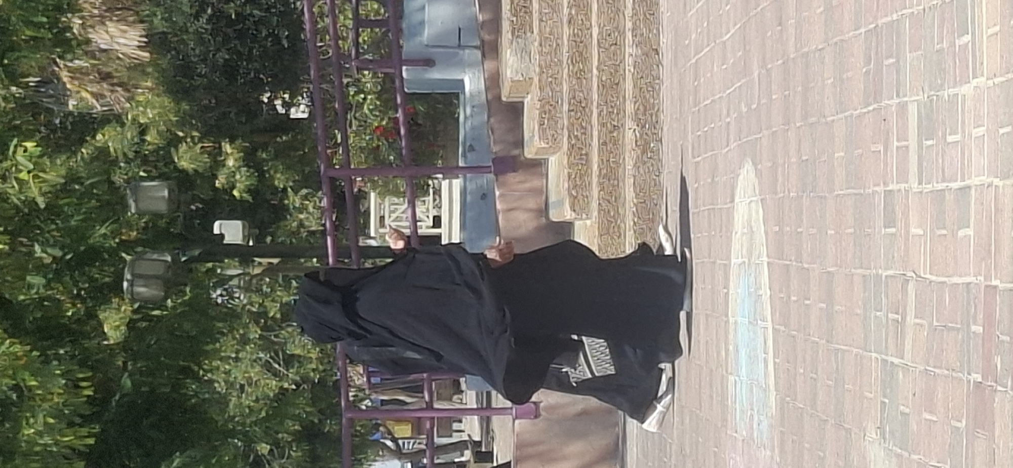 In Yerocham, Israel, a person walks on a paved pathway in front of a stone staircase. The person is wearing a full black abaya and hijab, with white sneakers. A purple railing runs along the side of the pathway, behind which are trees and decorative streetlights. The pathway is made of red bricks with lighter colored square inlays. The stairs are made of roughly cut, light-colored stones. The sky is visible through the leaves of the trees.
