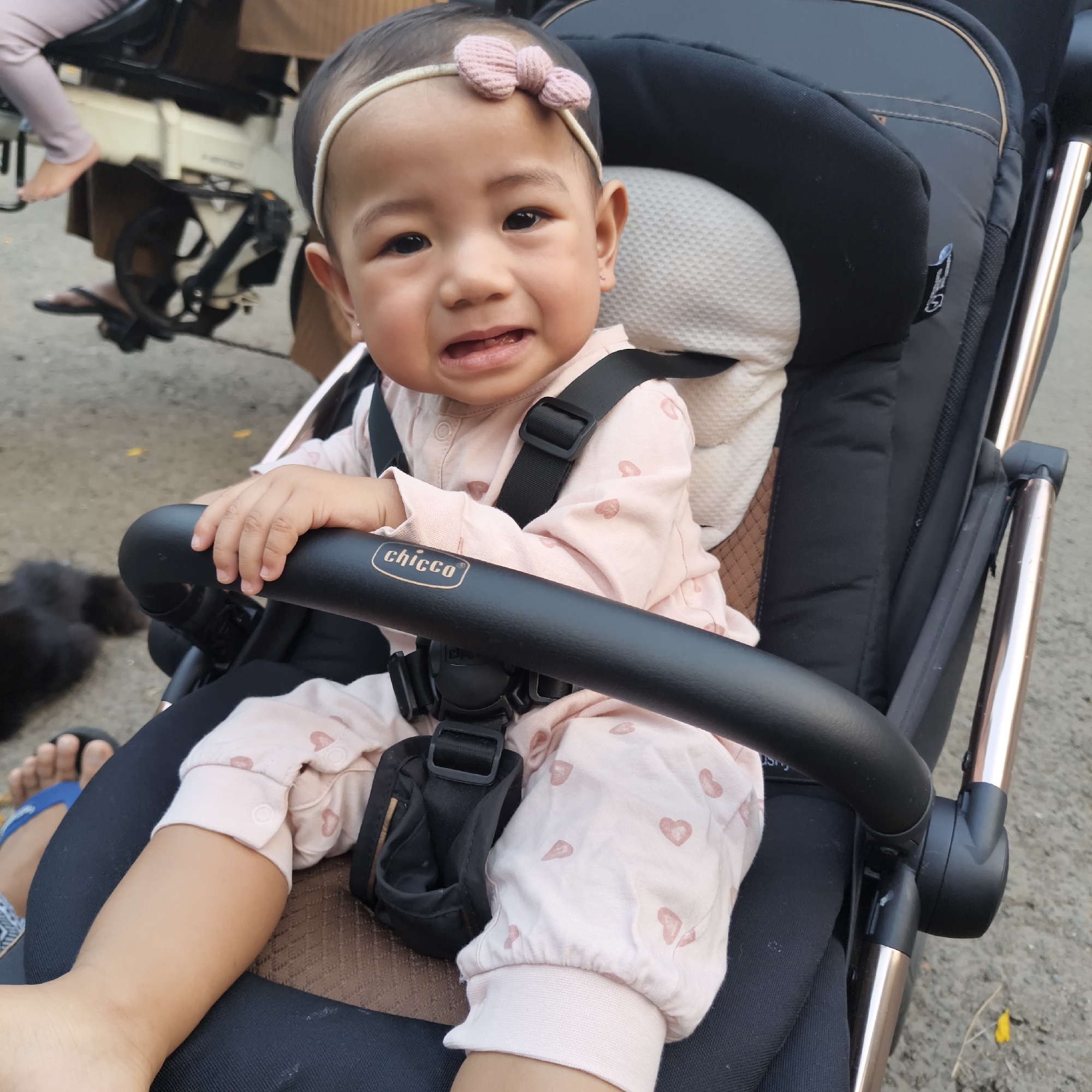 The image shows a close-up of a baby in a stroller. The baby is wearing a pink onesie with small heart patterns and a pink bow headband. The baby has its mouth slightly open and appears to be crying. The stroller has a black handlebar with the "Chicco" logo visible. The background is out of focus but appears to be an outdoor environment with pavement and some greenery. The scene suggests a public space, potentially a park or street in South Jakarta, Indonesia, given the context of the question.