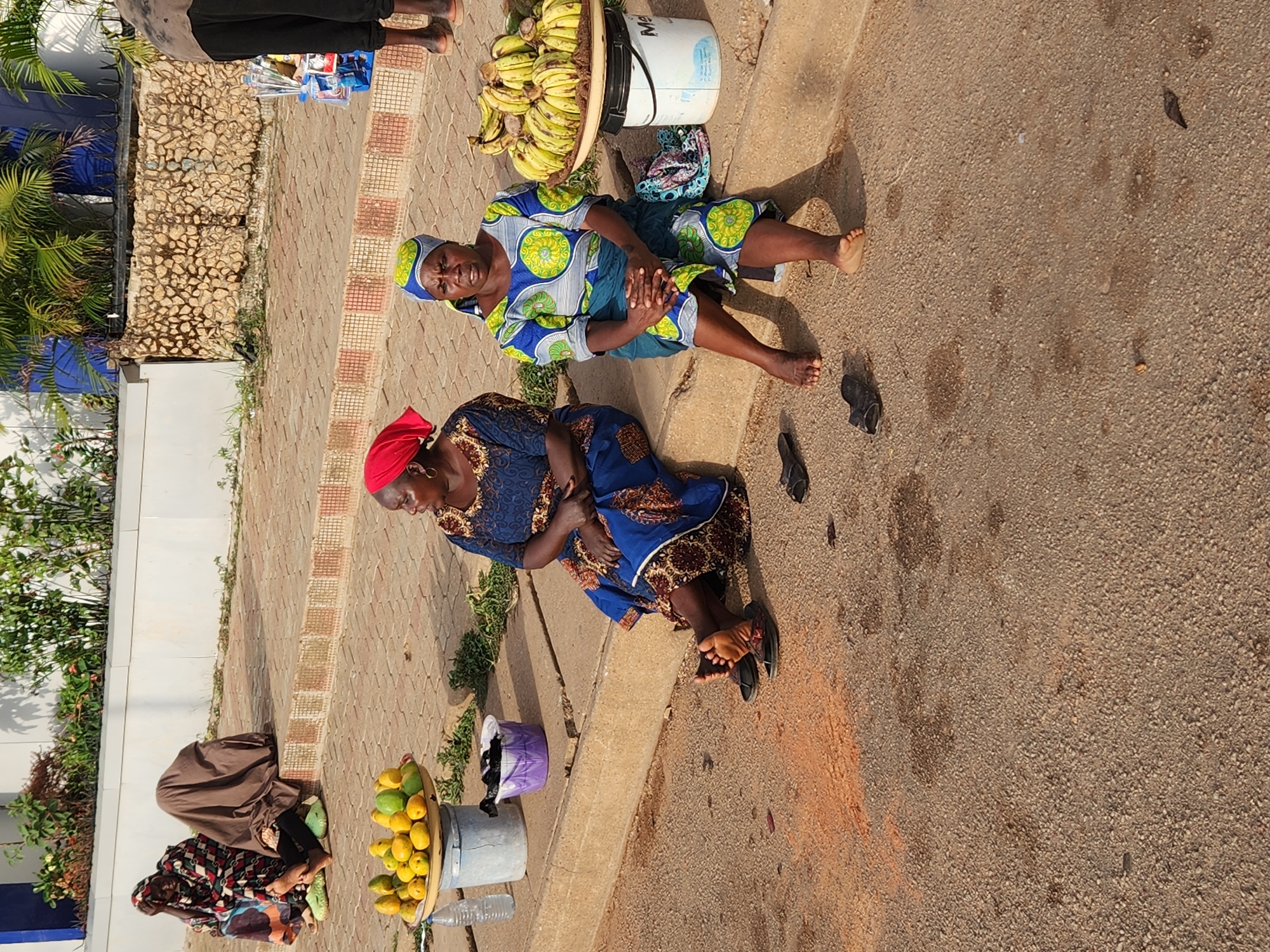 The image shows an outdoor scene in the city of Unknown, Unknown, with multiple women sitting on the side of a street selling goods. Two women are seated with a bucket of bananas between them. Two other women are seated with a bucket of mangoes. The women are dressed in colorful traditional clothing. One woman has a red head covering, while another has a blue and green head covering. Some of the women are barefoot. The background includes a low wall, vegetation, and a paved walkway. The street surface is paved. The lighting is bright, indicating it's daytime.