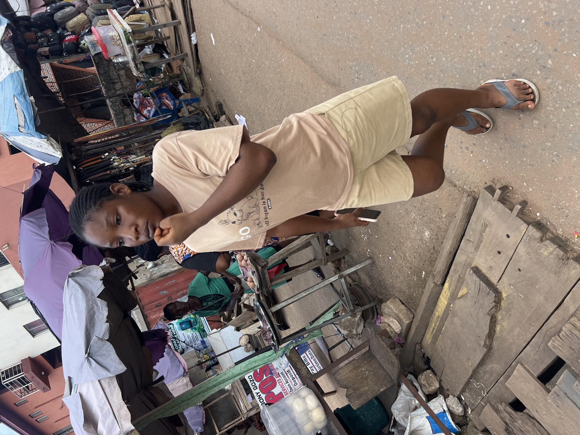This media file captures a street scene in Nkpor, Nigeria, with a focus on a young woman in the foreground. She is wearing a light brown t-shirt and beige shorts and is in the process of eating something. Her expression is thoughtful as she looks towards the camera. Behind her, the environment suggests a bustling market or roadside stall area.
Various objects are visible, including an assortment of bags, umbrellas, and what appear to be stalls with goods displayed. One stall has a sign with Nigerian text, including "BOSTADAKPOR," "OPAY," "POS," and other service-related terms like "Open Account," "Deposit," and "Pay Bills." Another sign reads "BONY GOZO." There are also stacks of wooden planks and scattered debris, typical of a less formal commercial area. The ground is a dusty, paved surface, and the lighting suggests it is daytime. The overall impression is of a lively, everyday scene in a Nigerian community.
