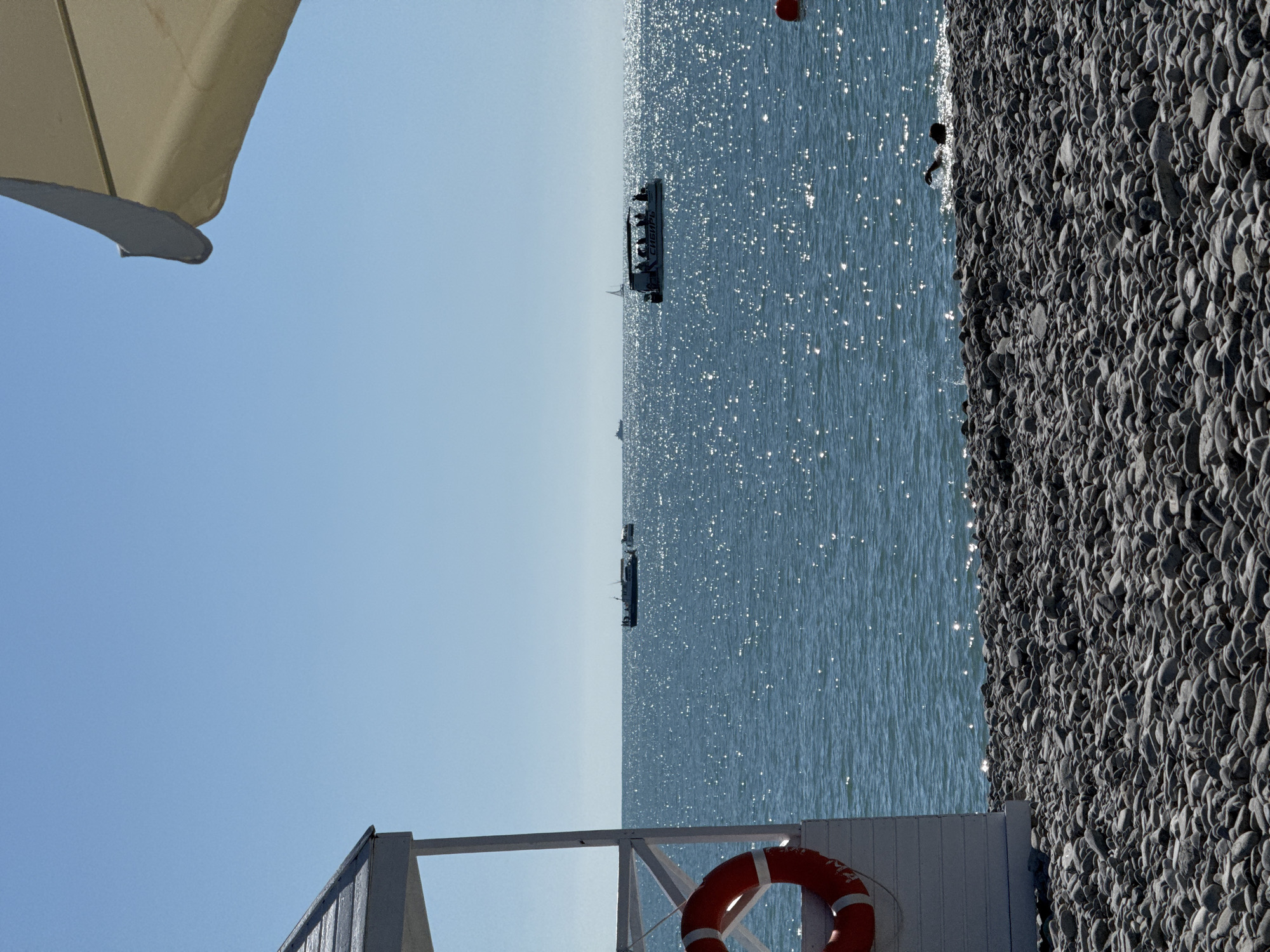 This image depicts a sunny day on a pebble beach in Sochi, Russia, looking out over a calm, sparkling sea under a clear blue sky.
In the foreground, the beach is composed entirely of dark grey pebbles of varying sizes. To the left, a portion of a white wooden structure is visible, likely a lifeguard station or changing cabin, with a red and white lifebuoy hanging on its side. In the upper right corner, the edge of a large, beige beach umbrella is partially visible.
The midground is dominated by the Black Sea, which exhibits a deep blue hue. The surface of the water intensely reflects the sunlight, creating numerous bright, shimmering points. Several boats are present on the water. Closer to the right-center, a motorboat with a canopy is seen in silhouette, carrying multiple people. The word "СНЕИРЦ" (SNIEIRTS or SNIEURTS) is partially visible on the side of this boat, likely a transliteration of "Сибирь" (Siberia). Further in the distance, closer to the horizon line, several other boats, including what appears to be a larger vessel and smaller craft, are visible. A small sailboat is also faintly discernible in the far distance. A red buoy floats in the water near the right side of the frame. Closer to the shore, on the right, a person is partially submerged in the water, actively swimming. The horizon line is distinct and straight.
