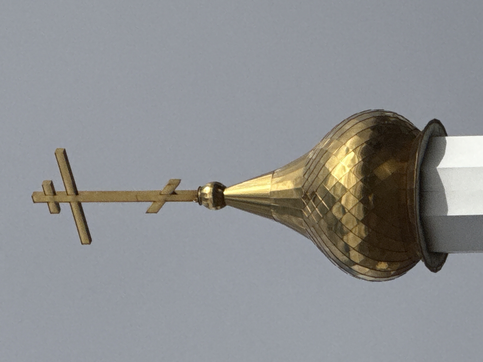 The image shows a close-up of the top of an Orthodox church dome against a gray sky. The dome is golden and onion-shaped, with a cross featuring three horizontal bars attached to its apex. The structure of the church that supports the dome appears to be white. It is impossible to verify this is the location in the city of Chaikovsky, Russia.