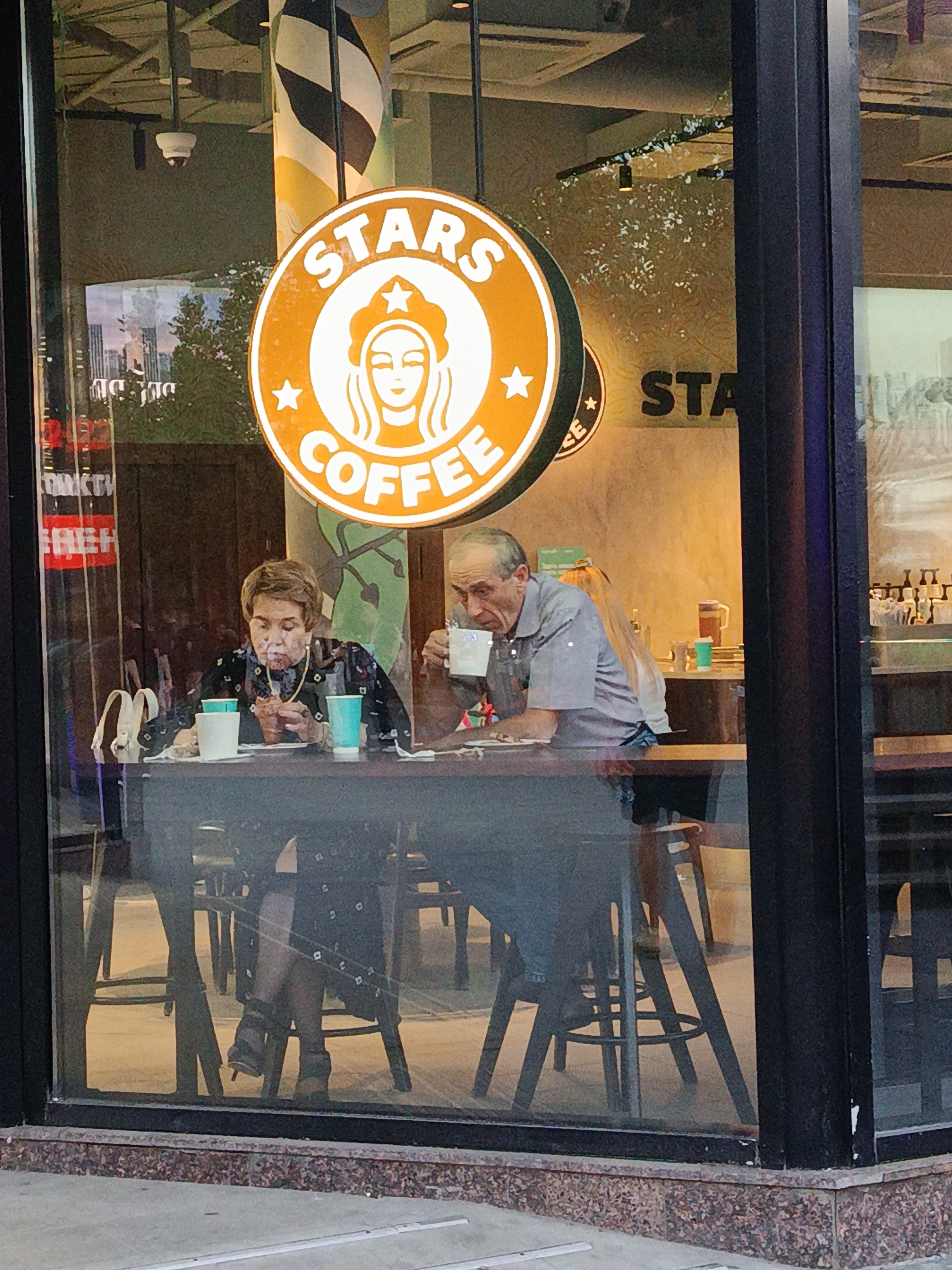 The image captures a view into a "Stars Coffee" establishment, likely located in Moscow, Russia, through a large glass window that exhibits reflections of the exterior environment.
In the foreground, a large circular orange and white "STARS COFFEE" sign is prominently displayed, featuring a stylized female face with a star on her head, encircled by text. To the left of this sign, a security camera is visible on the ceiling.
Two elderly individuals are seated at a dark wooden table inside. On the left, an elderly woman with short brown hair is wearing a dark, patterned long-sleeved top and a gold-colored necklace. She is looking down, seemingly interacting with an item in her hands, and appears to be using a nasal cannula for oxygen, with tubing visible around her ears and under her nose. In front of her are a white cup and two light blue/teal cups. Her feet, clad in dark sandals with a low heel, are visible beneath the table.
Across from her on the right, an elderly man with receding hairline and some grey hair is wearing a light blue/grey short-sleeved collared shirt. He holds a white cup to his mouth, drinking from it, and looks towards the left. A lighter-haired individual is partially visible behind him.
The interior background reveals more tables and stools, another sign partially obscured but showing "STA," and a black and white striped pillar. Reflections on the glass include greenery, faint outlines of distant buildings, and a partial white sign with red text on the far left. The bottom of the window is set into a speckled red and brown granite-like base, with a grey concrete sidewalk visible in the immediate foreground.
