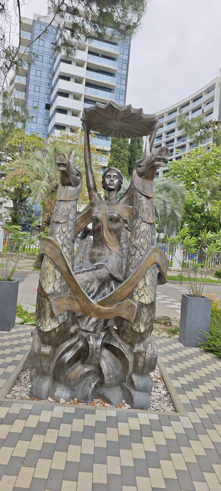 The image depicts a statue in Sochi, Russia. The statue features a central female figure holding a large lotus leaf above her head, flanked by two dragon-like figures. The statue is made of a bronze-colored material. The statue is located on a paved area with gray and beige bricks. In the background, there are buildings, trees, and other vegetation. Two rectangular planters are also visible. The sky is overcast.