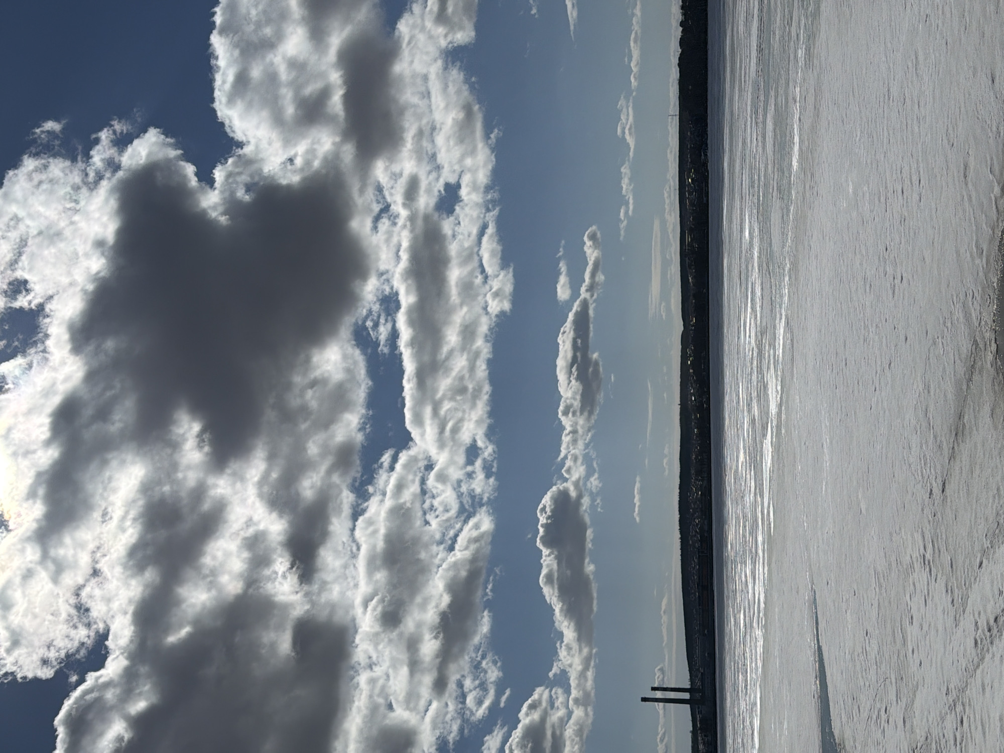 The image depicts a winter scene in Votkinsk, Russia. A frozen body of water (likely the Votkinsk Reservoir) occupies the right portion of the image. The water surface is covered in snow and ice, reflecting sunlight. Along the distant shoreline, a thin line of trees and buildings is visible. Two smokestacks are seen rising above the treeline. The sky, taking up the left portion of the image, features a mix of blue sky and various cloud formations. Some clouds are dark and dense, while others are lighter and more scattered.