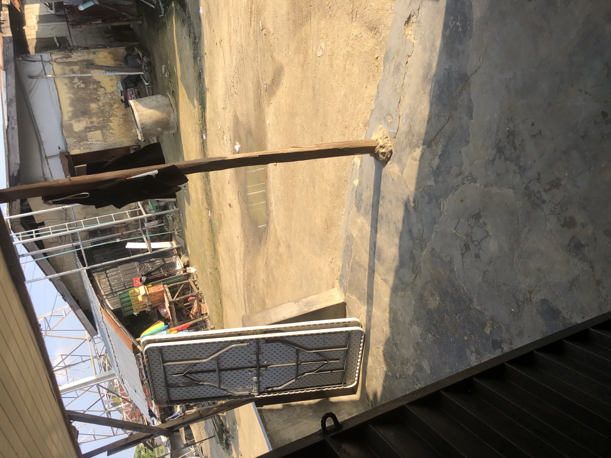 The image depicts an outdoor scene in a potentially urban or semi-urban environment in Ado, Nigeria. In the foreground, a concrete surface is visible, with a dark, ribbed structure to the left, possibly part of a staircase or seating area. A large, folded table with a grey, patterned surface stands upright against a concrete barrier.
The middle ground shows a dirt or sandy ground, with a small puddle of water and scattered debris. A prominent wooden post, with rough, earthy material around its base, stands vertically in this area. Beyond the post, a modest structure with a corrugated metal roof is visible, appearing to be a shop or stall. Shelves with various items, including boxes and possibly food products, are stacked outside this structure. A ladder is leaning against another building in the background.
Further back, a well-like concrete structure is present, and more buildings with weathered walls and corrugated roofs are visible. The overall impression is one of a bustling, informal marketplace or residential area, possibly characterized by resourcefulness and a somewhat unkempt environment. The lighting suggests it is daytime, with shadows indicating the sun's position. No people are clearly visible or interacting within the frame.