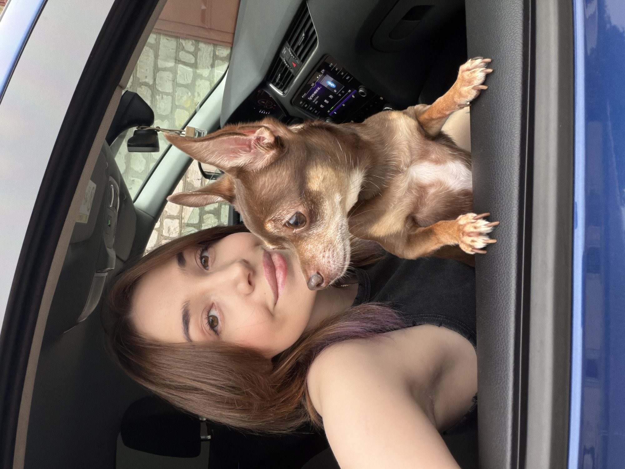 A young woman with brown hair and a black tank top is in the driver's seat of a blue car, with her arm resting on the open window frame. A small, brown chihuahua-mix dog is next to her, also resting its front paws on the window frame. The dog is looking to the right, while the woman is looking forward, smiling. The car's interior is visible, including the dashboard and a radio. The scene appears to be outdoors, with a brick wall in the background. The location is unknown.