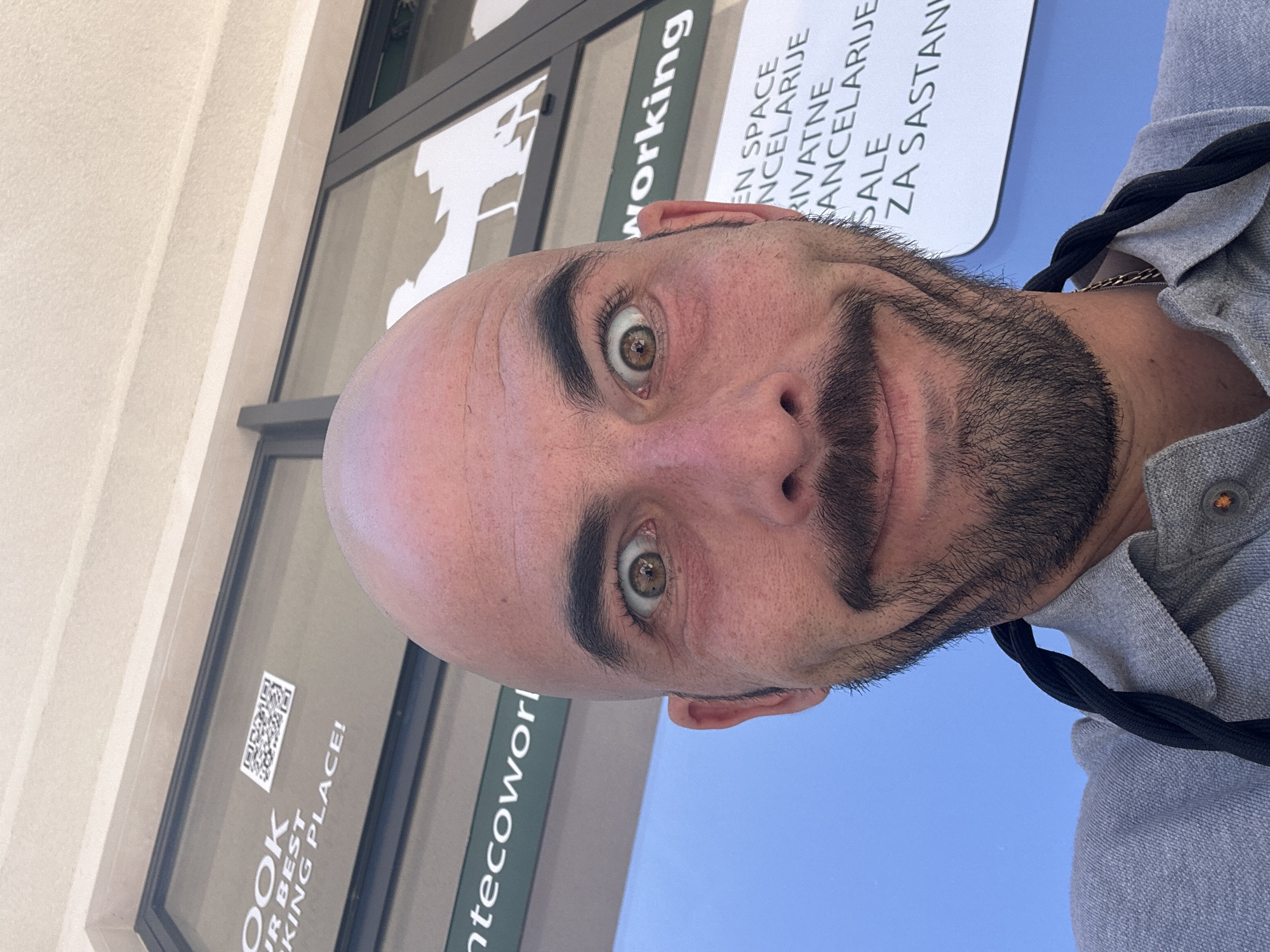The image is a close-up selfie of a bald, bearded man in front of a business establishment. The man is fair-skinned with hazel eyes. He is wearing a grey shirt with a black lanyard around his neck.
Behind the man is a storefront window and signage. The sign includes the partial words "working," "OPEN SPACE," "ANCELARIJE," "PRIVATNE," "CANCELARIJE," "SALE," and "ZA SASTAN." Another sign displays "OOK" and "JR BEST KING PLACE!" as well as "ntecoworl." A QR code is also present in the window. Based on the text on the sign, specifically "ANCELARIJE," "PRIVATNE," and "ZA SASTAN," the location is likely in Montenegro, possibly Budva. The signage suggests it is a co-working space offering office rentals.