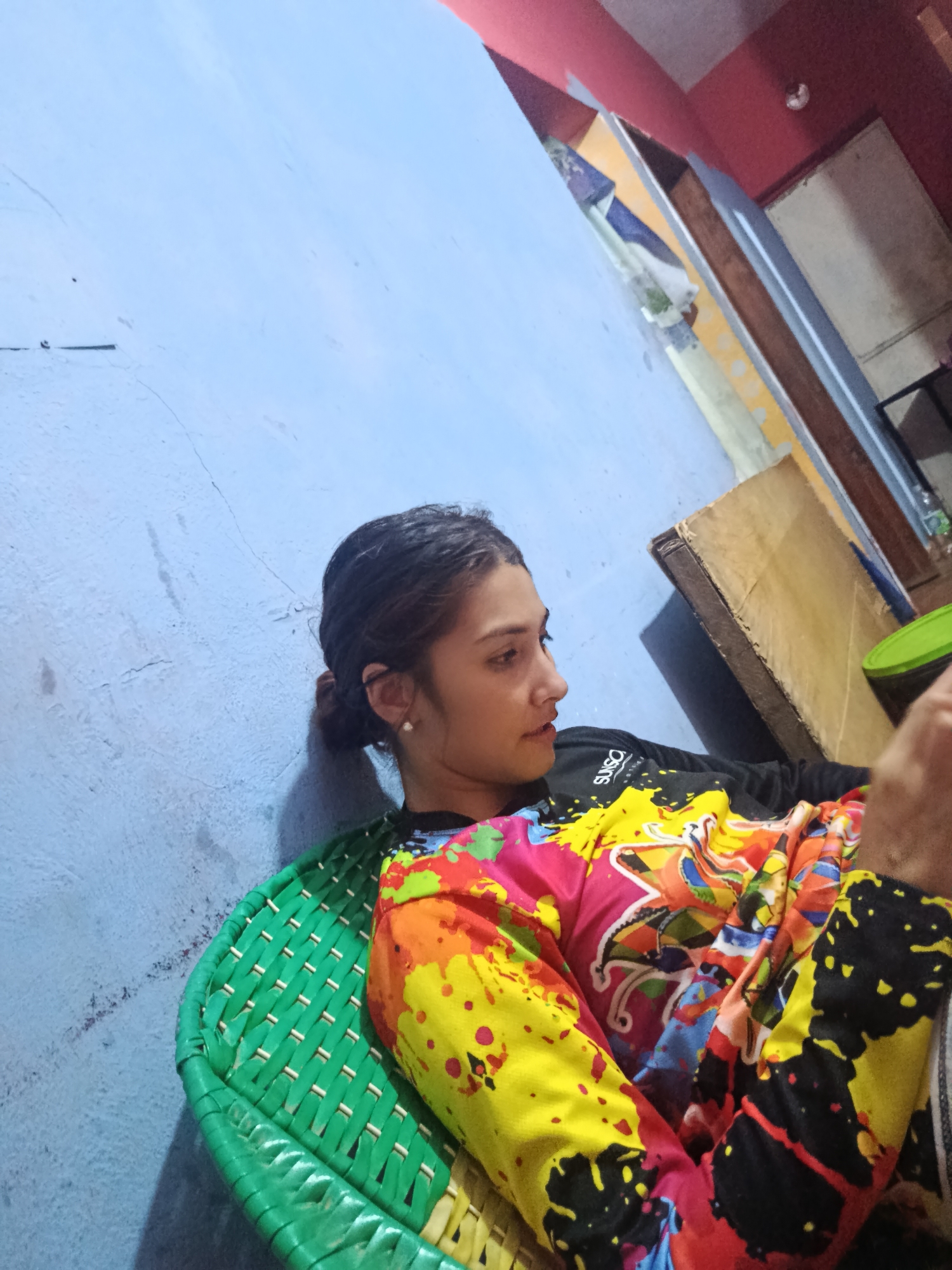 A person sits on a green woven chair in a room. The walls are painted blue and red. The person has their dark hair pulled back in a low bun and is wearing a colorful shirt with splatter paint designs over a black long-sleeve shirt. A cardboard box sits on the floor near a doorway. This image was likely taken in Juan Griego, Venezuela.