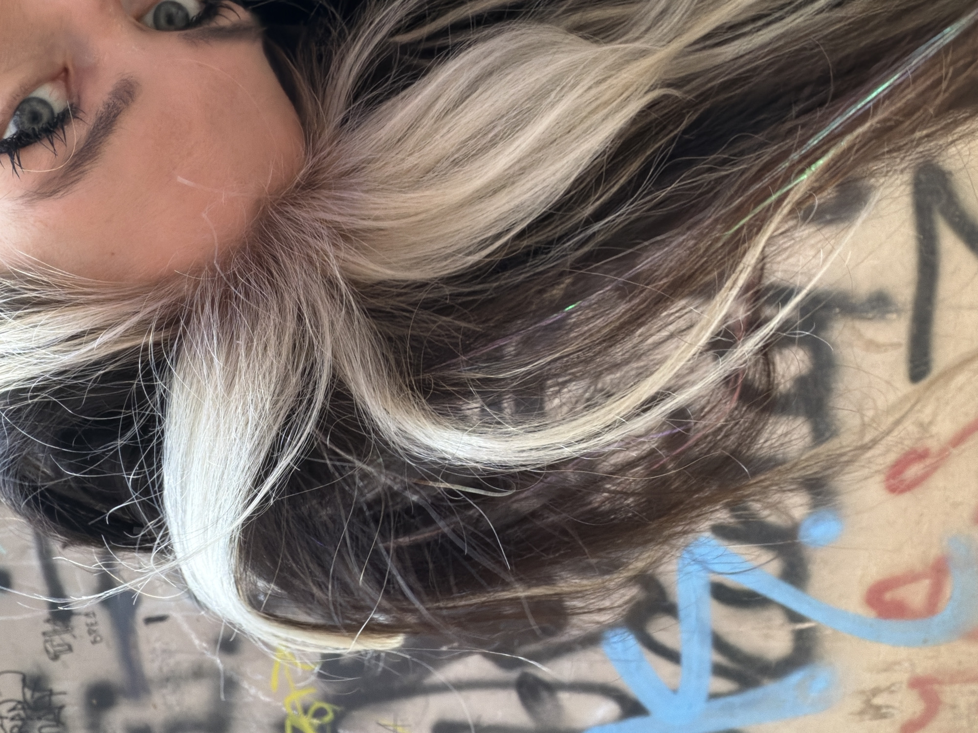 A close-up, high-angle shot captures the upper left portion of a person's face and their voluminous hair, set against a graffiti-covered surface. The person's left eye is prominently visible, featuring a striking blue-green iris with dark, defined eyebrows and mascara-laden eyelashes. Their skin appears fair.
The hair is long and flowing, presenting a vibrant contrast between dark brown strands and numerous thick, bright blonde or silver highlights. A distinctive detail within the darker hair is a single thin, iridescent strand that shimmers with faint hues of light green and pink, suggesting a hair tinsel.
The background is a textured, light-colored surface, likely a wall or ground, extensively adorned with spray-painted graffiti. This graffiti includes abstract shapes, bold lines, and partial letters in various colors such as black, light blue, red, and yellow. Elements like broad blue lines and curved red shapes are discernible, along with darker, intricate scribbles.
The lighting is bright and natural, indicating an outdoor setting. The person appears to be lying down or reclining, gazing slightly upwards towards the camera. No specific activity is occurring beyond this pose. While various graffiti marks are present, no clearly legible full words are discernible within the visible text. The overall scene has an urban, artistic aesthetic.