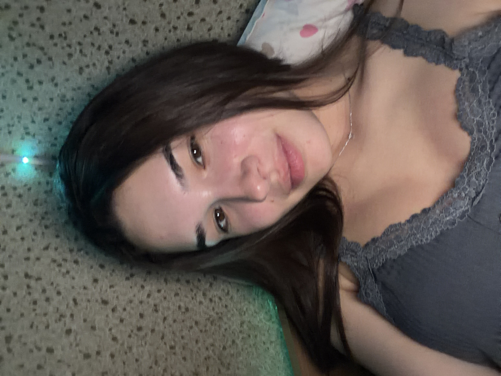 A young woman with dark hair lies on a surface, possibly a bed or the floor, with her head resting on a pillow. She is wearing a gray top with lace detailing around the neckline. A thin silver necklace is visible on her neck. Her skin appears clear and well-lit. To the left of her head is a textured surface. The environment is illuminated by a light source, possibly a neon light, which emits a greenish-blue glow near the edge of the image. The location, Sofia, Bulgaria, is not discernible from the provided image.