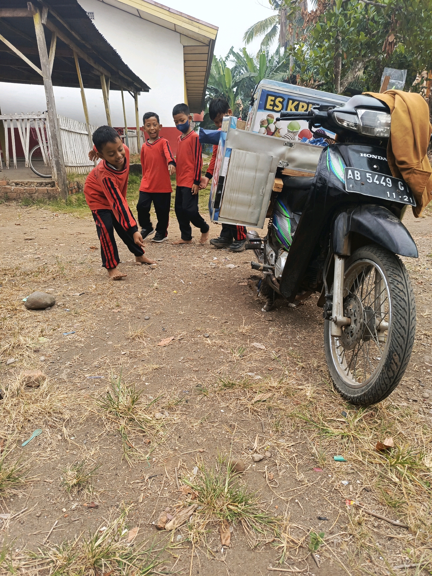 In Megang Sakti II, Indonesia, four young boys in red shirts and black pants stand around an ice cream cart attached to a motorcycle. The boy closest to the camera, barefoot, is looking down at the ground and appears to be playing with a stone. The other three boys are looking towards the ice cream cart, with one boy wearing a blue mask. The motorcycle, a Honda, has a license plate reading "AB 5449 G 11-21". The background shows a white building with a wooden overhang and a white fence. The ground is dirt and dry grass, with some scattered rocks and debris. A brown jacket is draped over the back of the motorcycle.