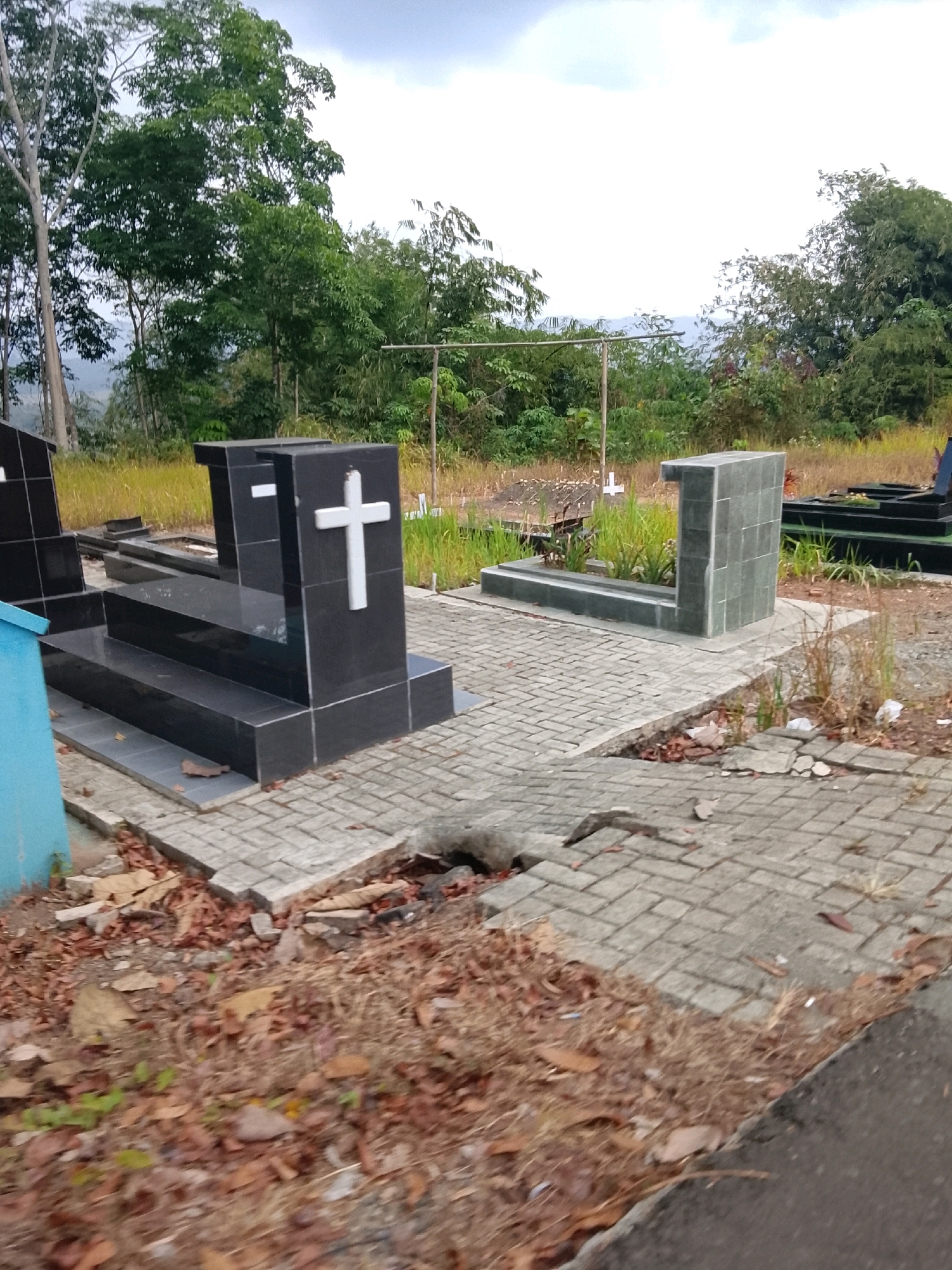 The image depicts a cemetery in Banjar, Indonesia. In the foreground, a grave marker made of polished black stone with a white cross is visible. The marker has a stepped base. To the right, another grave marker constructed with gray tiles is seen, with a low rectangular planter box in front. The cemetery is paved with interlocking bricks, some of which are cracked or displaced, with dried leaves and debris scattered on the ground and between the bricks. Beyond the graves, dense green trees and vegetation form the background, with hills visible in the distance. The sky is overcast with white clouds. The scene is quiet and appears to be an outdoor daytime shot.