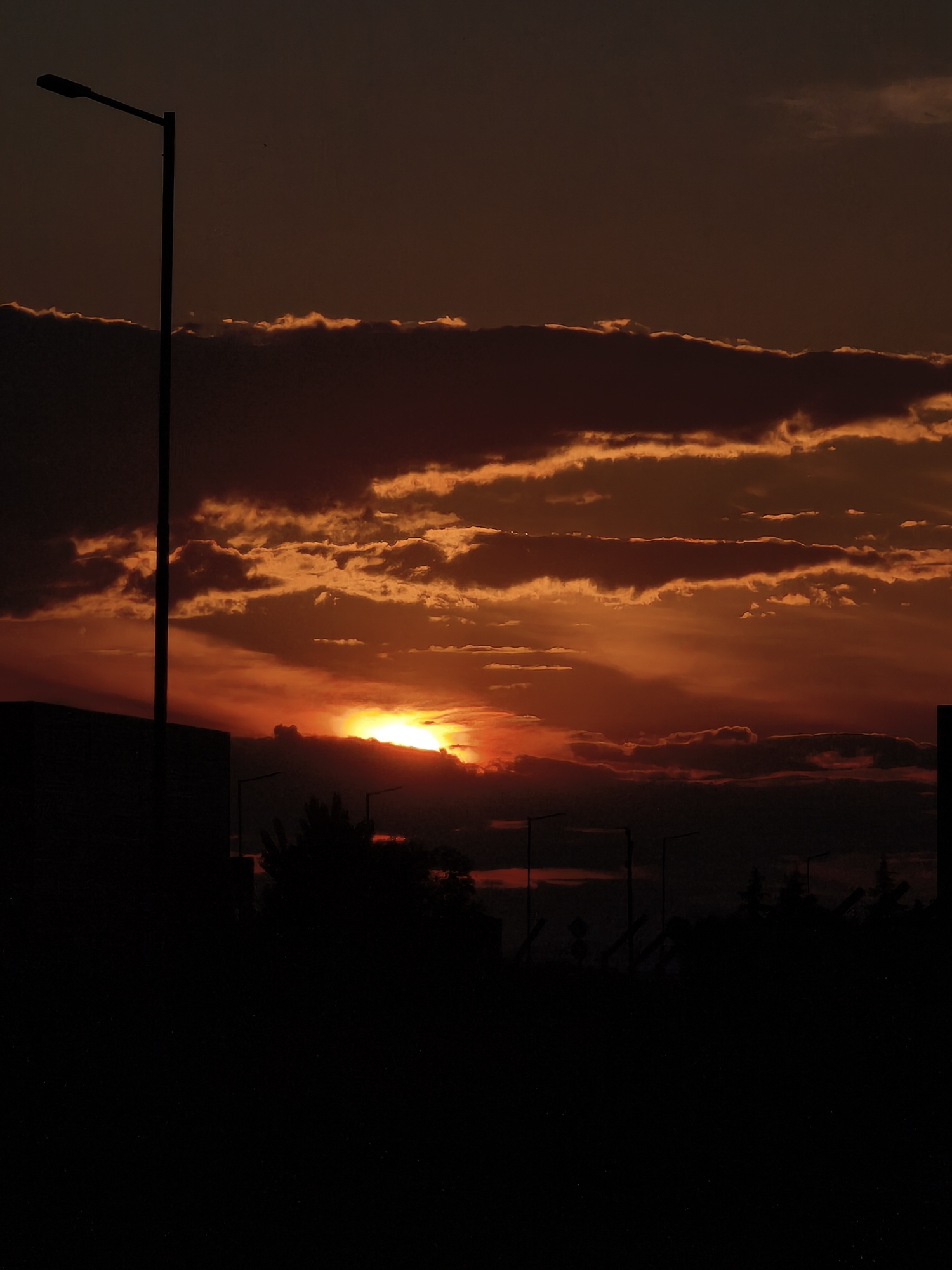 A vertical shot depicts a fiery sunset in what is likely the city of Benkovski, Bulgaria. The sky is streaked with horizontal bands of dark clouds illuminated by the setting sun, creating a fiery orange hue. A silhouetted lamppost stands on the left. The horizon features the silhouettes of buildings, trees, and additional lampposts. The bottom of the frame is dominated by deep black.
