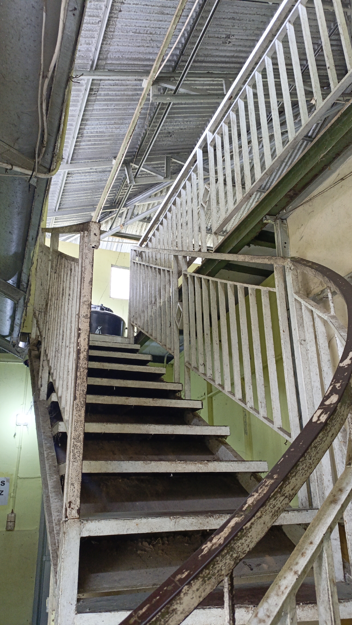 The image shows an industrial-style spiral staircase made of rusted metal. The staircase has a white railing that is also showing signs of rust and wear. The steps are made of metal and appear to be dark in color. The staircase is located indoors, under a corrugated metal roof. There is a window visible at the top of the staircase, letting in some natural light. In the background, there is a large black tank. The walls appear to be painted a pale yellow. This scene is located in the city of Trihanggo, Indonesia.