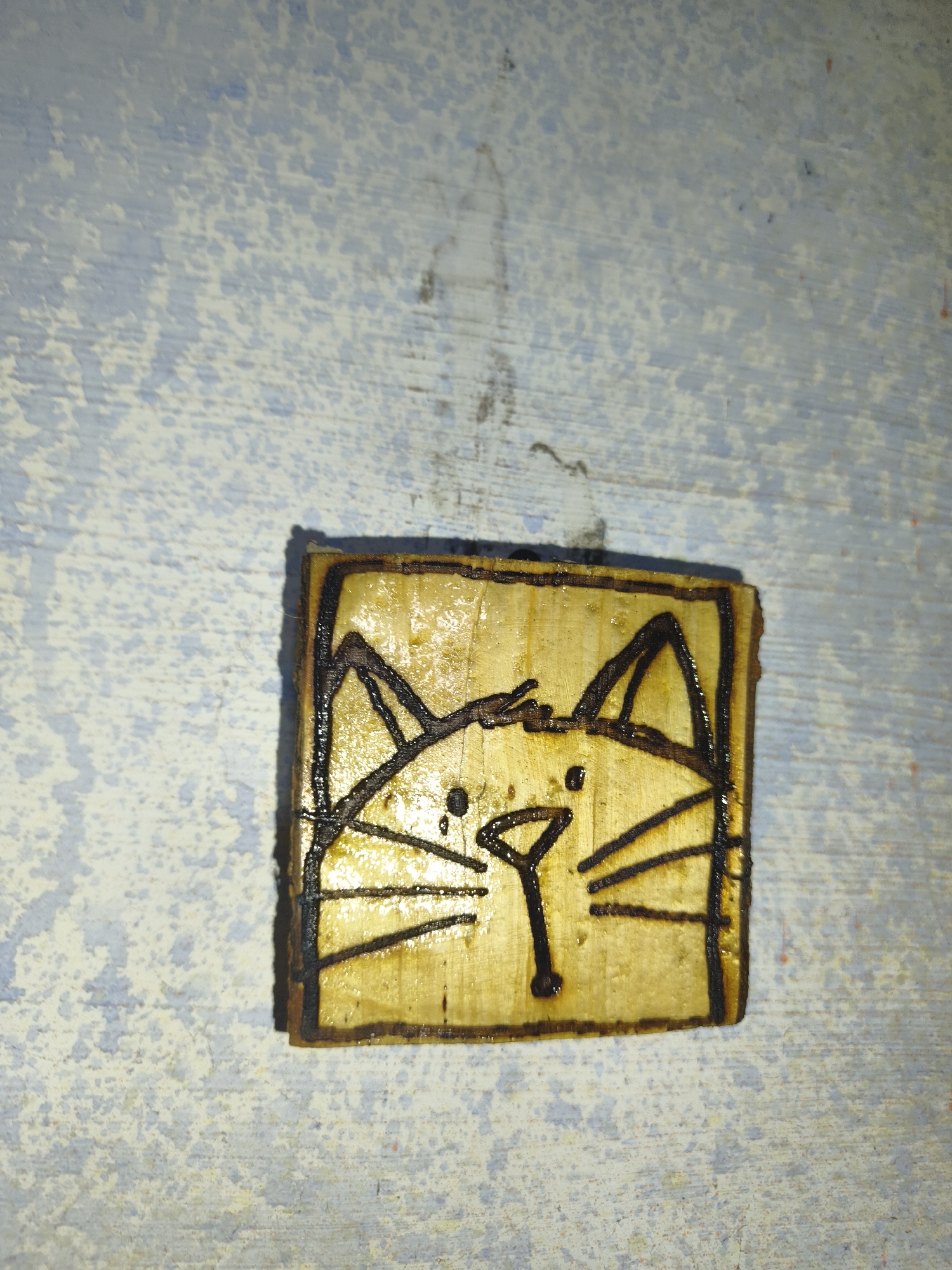 A small, square piece of wood, approximately 2x2 inches, is the central focus of the image. The wood has a light, golden-brown color and appears to be treated with a sealant, giving it a slightly glossy finish. A simple drawing of a cat's face is etched or burned into the wood. The cat's features are rendered with thick, dark lines. Two triangular ears sit atop its head, and two almond-shaped eyes are visible. A small, inverted triangle forms the nose, with a dot below it representing the mouth. Several straight lines extending outwards from the nose suggest whiskers. The edges of the wood are bordered by darker, burned lines. The wood object is positioned on a surface with a distressed, textured appearance. This surface is predominantly a light, off-white color with irregular splotches and streaks of pale blue and gray. The texture suggests a painted or weathered material, possibly wood or plaster. The lighting in the image creates a subtle shadow cast by the wood object to its lower left. The overall impression is that of a handcrafted item, perhaps a small token or decorative element, placed on a rustic surface. The location is in Гаранскі сельскі Савет, Belarus.