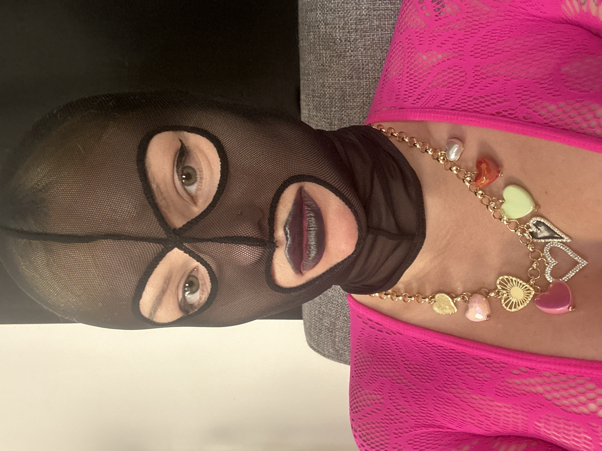 A woman wearing a black fishnet balaclava and bright pink fishnet top is positioned indoors. She is also wearing a gold necklace with heart-shaped and pearl charms. The background consists of a cream-colored wall and a gray textured surface.
There is no specific information about this picture being taken in the city of Budva, Montenegro, therefore the exact location cannot be provided.