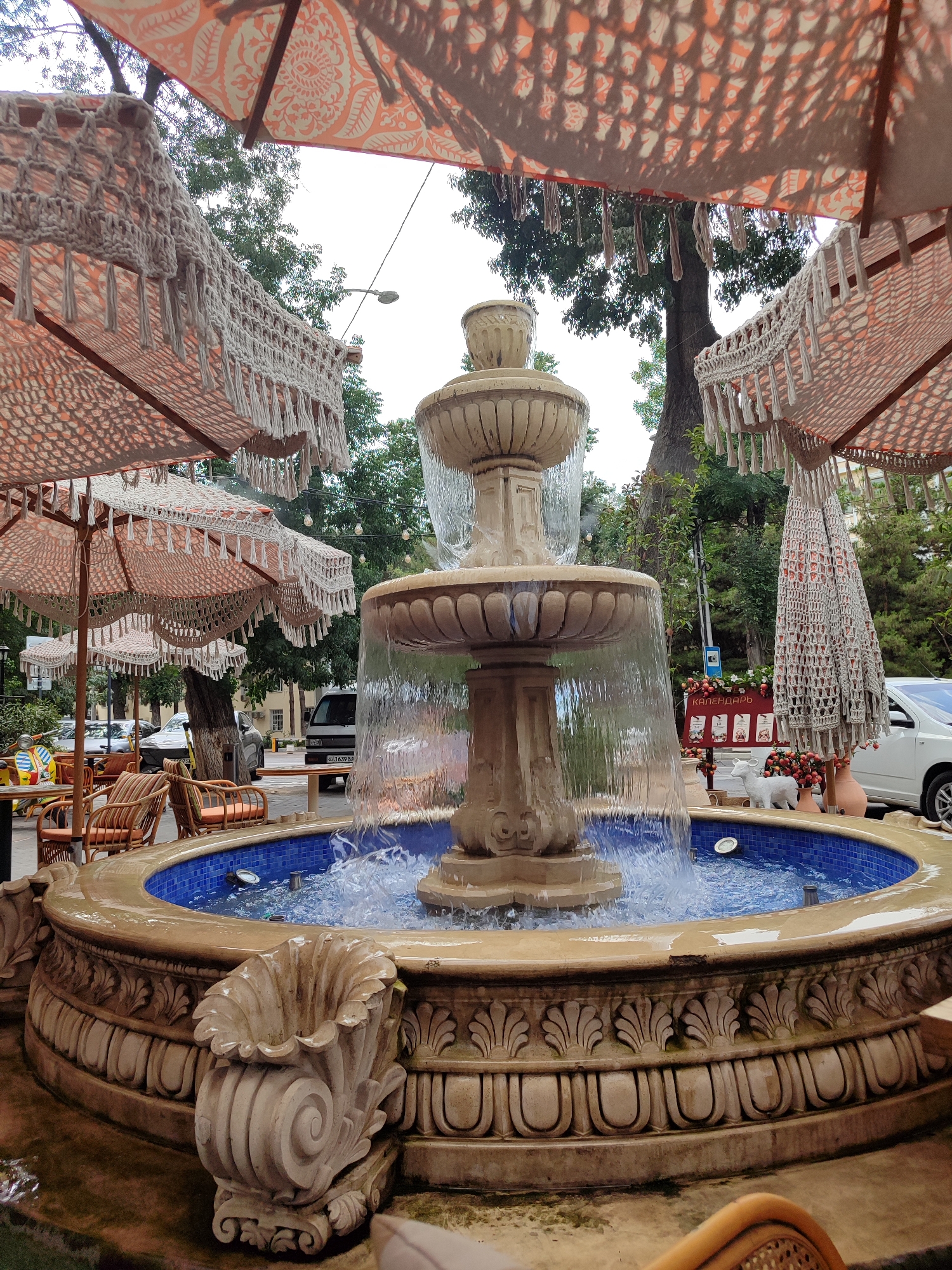 The image depicts a multi-tiered stone fountain with water flowing over its edges into a blue-tiled basin. The fountain is situated outdoors, likely in a courtyard or public space, surrounded by decorative umbrellas with macrame fringe. In the background, trees, buildings, and parked vehicles are visible, suggesting an urban environment. The overall ambiance suggests a cafe or restaurant setting, given the presence of outdoor seating, including wicker chairs and tables with orange cushions. A sign in Russian that reads "КАЛЕНДАРЬ" (Calendar) is also visible, along with decorative items like a white dog figurine and potted plants. The specific location is identified as Tashkent, Uzbekistan.