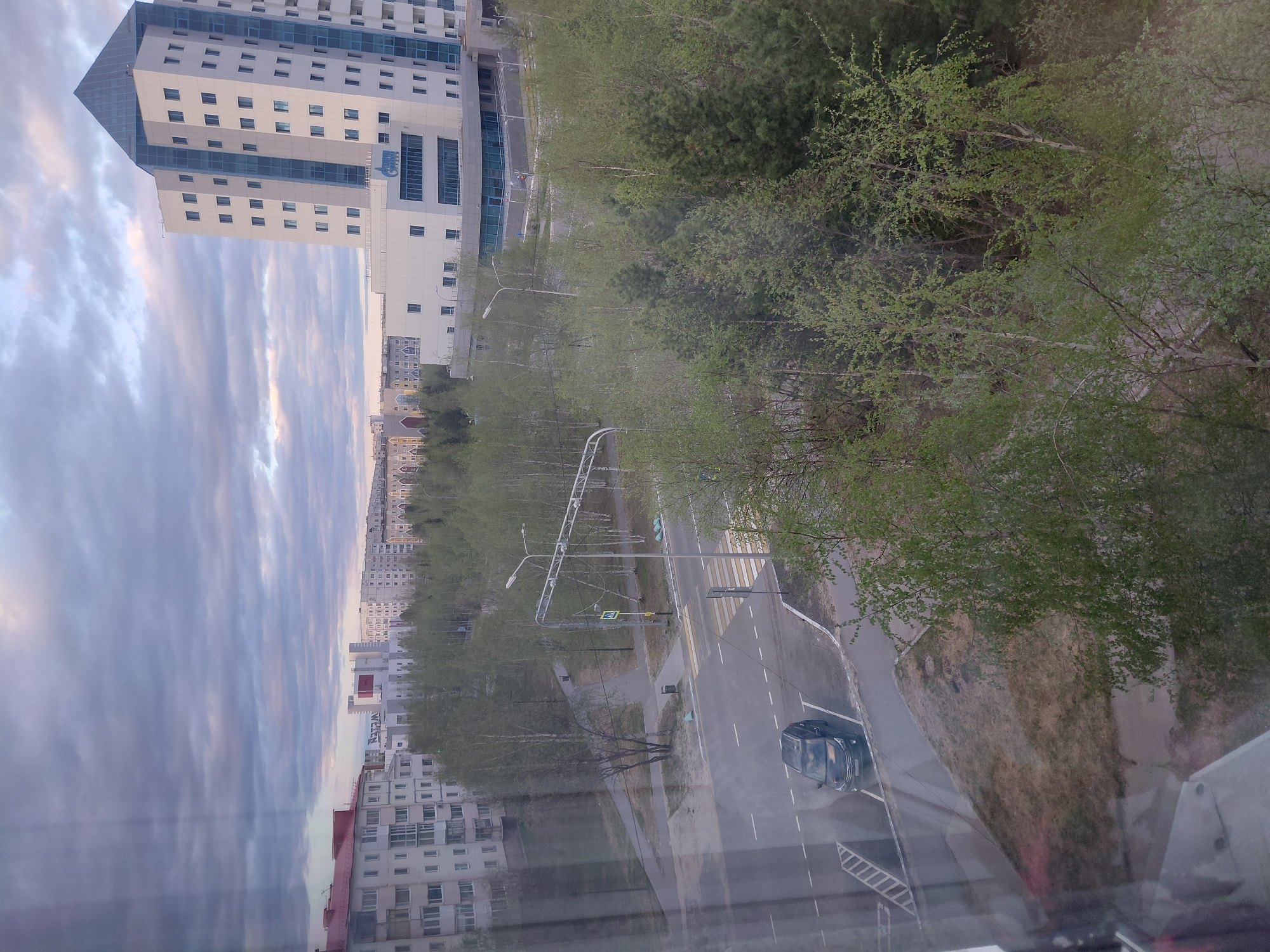 This image captures an urban street scene in Nadym, Russia, from an elevated perspective, likely a window, indicated by reflections at the bottom. The sky is overcast with long, soft cloud formations, suggesting either early morning or late afternoon light.
The prominent subject is a tall, modern building on the left (when correctly oriented), featuring a distinctive angled, multi-faceted roofline and a facade of light-colored panels interspersed with horizontal bands of blue-tinted glass windows. On one of the blue glass sections, Cyrillic letters resembling "ОММ" are visible, likely a company or institutional name. Adjacent to this building and further down the street are other multi-story residential buildings.
A wide asphalt street runs through the middle ground, featuring clear white lane markings and a prominent pedestrian crosswalk with zebra stripes. A dark-colored car, possibly an SUV or hatchback, is visible on the street, either parked or moving slowly. Streetlights and traffic signals, including a visible green light, are present.
The street and surrounding areas are extensively lined with numerous deciduous trees, their branches adorned with fresh, vibrant green leaves, indicating the spring season. Some evergreen trees are also interspersed among them. Sidewalks and grassy areas separate the street from the buildings and tree lines. The overall scene depicts a quiet, modern urban environment during a cloudy spring day.