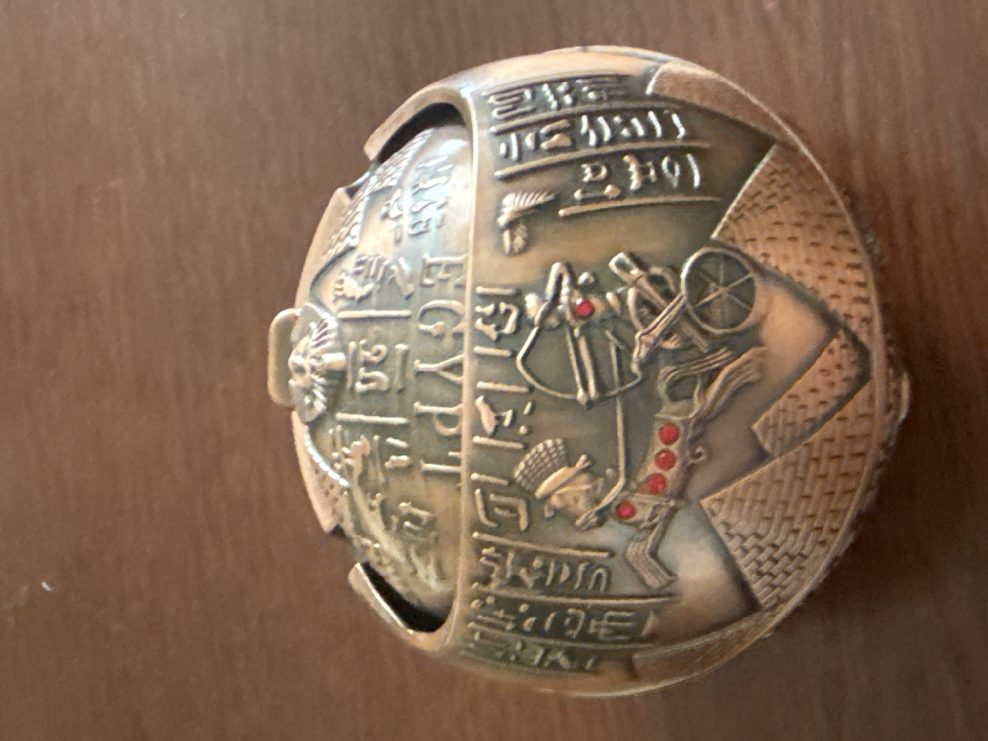 The image shows a round, decorative object with an Egyptian theme. The object is made of metal, possibly brass or bronze, and has an aged, antique appearance. It is adorned with intricate carvings and hieroglyphics. On the surface, there is a depiction of a chariot with a figure drawing a bow, pulled by a horse. The chariot wheels are visible, and the horse is decorated with red gemstones. Pyramidal shapes are etched at the base of the object, and rows of hieroglyphs are present on the lid and the sides. The object is placed on a brown, wooden surface, which appears to be a table. The lighting suggests an indoor setting with soft illumination. No specific location in a city is identifiable.