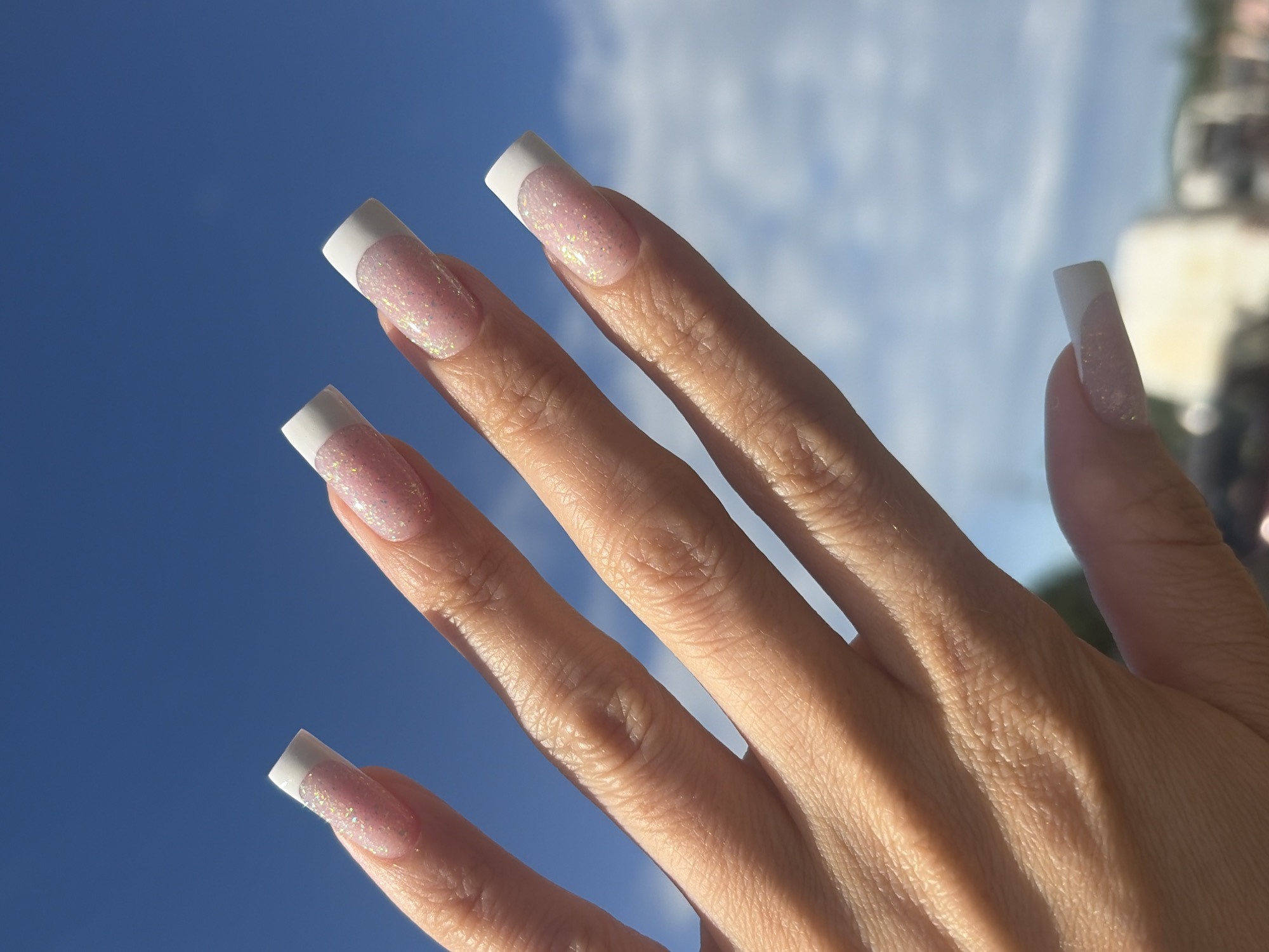 The image features a close-up view of a person's hand with manicured nails, set against a backdrop of a bright blue sky with wispy white clouds. The nails are long and square-shaped, with a classic French manicure style, featuring a white tip over a sheer, glittery pink base. The sunlight casts distinct shadows across the skin of the hand, highlighting the texture of the skin and the structure of the fingers. The overall impression is one of bright daylight, possibly late morning or early afternoon, given the strong illumination and clear sky. The upward angle of the shot suggests the hand is held up towards the sky. There are no discernible location cues specific to Saint Petersburg, Russia, beyond the general outdoor scene. The image does not contain any visible text.