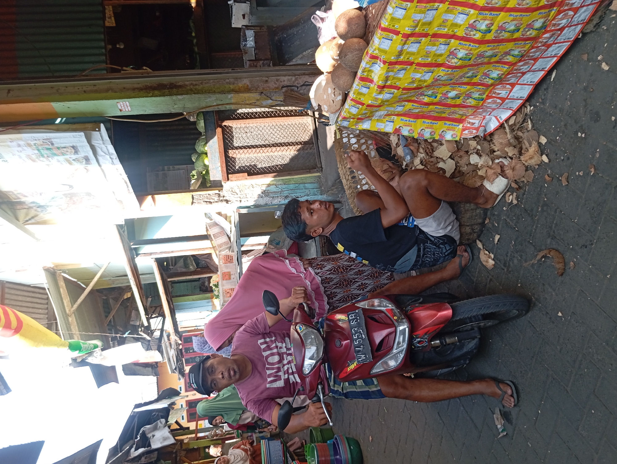 In the city of Kepulungan, Indonesia, a street market scene unfolds. A man wearing a maroon t-shirt and a black baseball cap sits on a red scooter, his left hand on the handlebars. He is accompanied by a woman in a pink headscarf and patterned skirt. To the right, a young man in a black t-shirt and gray shorts is seated on a wooden log, holding what appears to be a piece of coconut. He is surrounded by coconut husks and shells. In the background, stalls with various goods are visible, including baskets of produce and other market items. The ground is paved with dark gray tiles. The overall atmosphere suggests a bustling local market.
