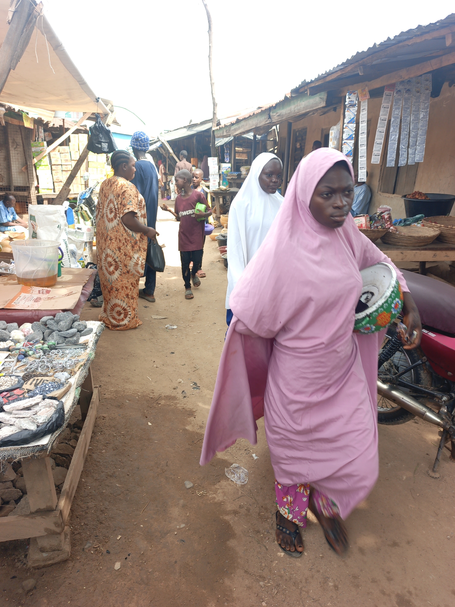 The image captures a bustling street scene in Akwanga, Nigeria. The setting is a market area with stalls lining both sides of a dirt road.
In the foreground, a woman wearing a light pink hijab and floral pants is walking towards the viewer, carrying a bowl. Behind her, another woman in a white hijab walks in the same direction. Further back in the scene, a woman in an orange patterned dress and a person wearing a blue hat stand near a vendor stall. A child in a maroon shirt is also visible, walking in the same direction as the women.
On the left side of the image, a vendor's stall displays various items, including stones and metal objects. On the right side, a motorcycle is parked near a stall selling hanging merchandise. The environment appears to be a busy market, with various people milling about.