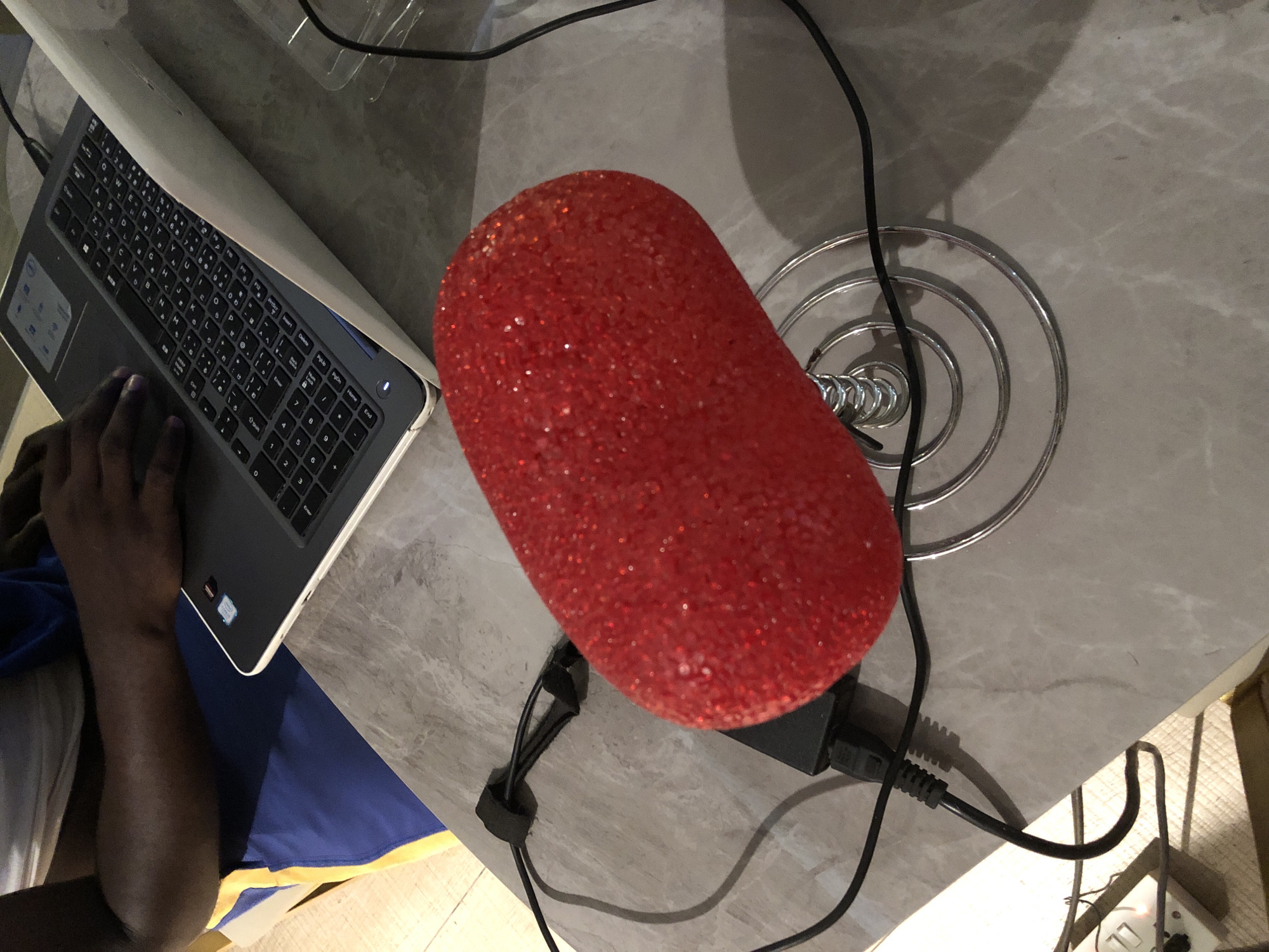 A high-angle shot depicts a laptop, a red, sparkling decorative lamp, and a partial view of a person sitting at a gray, possibly marble-topped table. The laptop is open, with a hand visible on the trackpad. To the right of the laptop, there's a red, oval-shaped lamp adorned with what appear to be small, glittering beads. The lamp is connected to a power source via a black cord and plug. A portion of a person's arm and torso is visible on the left side, clad in a white shirt and blue shorts. The scene seems to be set in a domestic or office environment. Location: Unknown, Unknown.